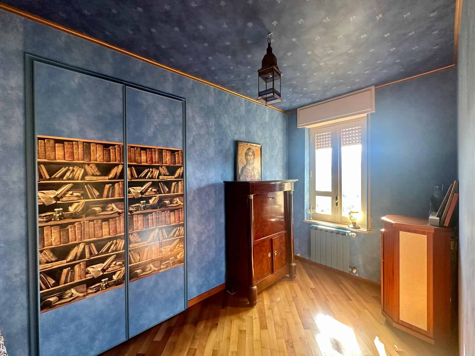 Armadio cabine con stampa libreria, pareti e soffitto blu, mobili in legno, finestra con tapparella, mobile antico, arredo classico, casa moderna, interior design, stile tradizionale, decorazioni eleganti, NextCasa.