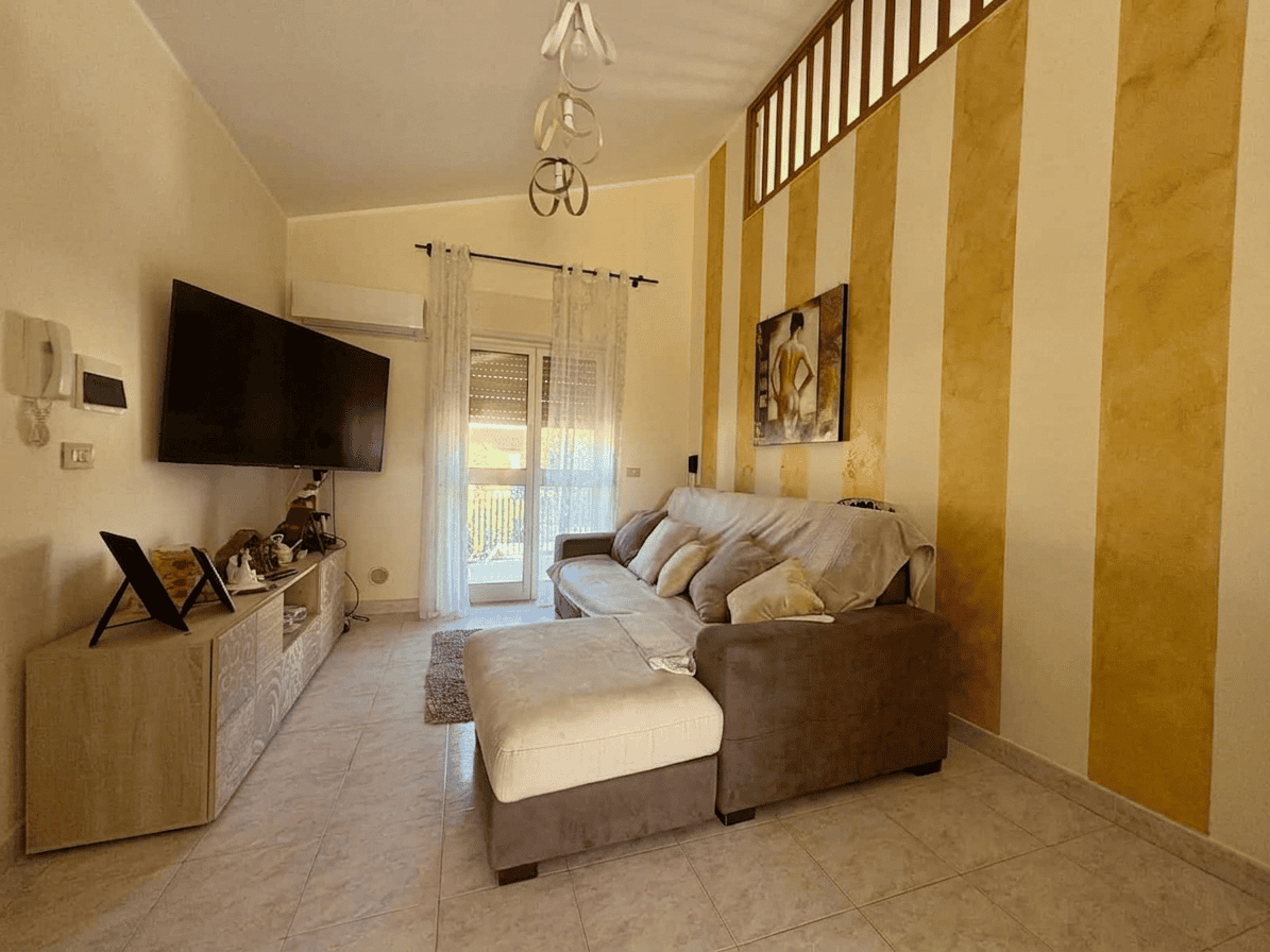 Interno di un soggiorno luminoso con divano, TV a parete e pareti decorate con strisce dorate, ideale per affitti turistici o residenziali.