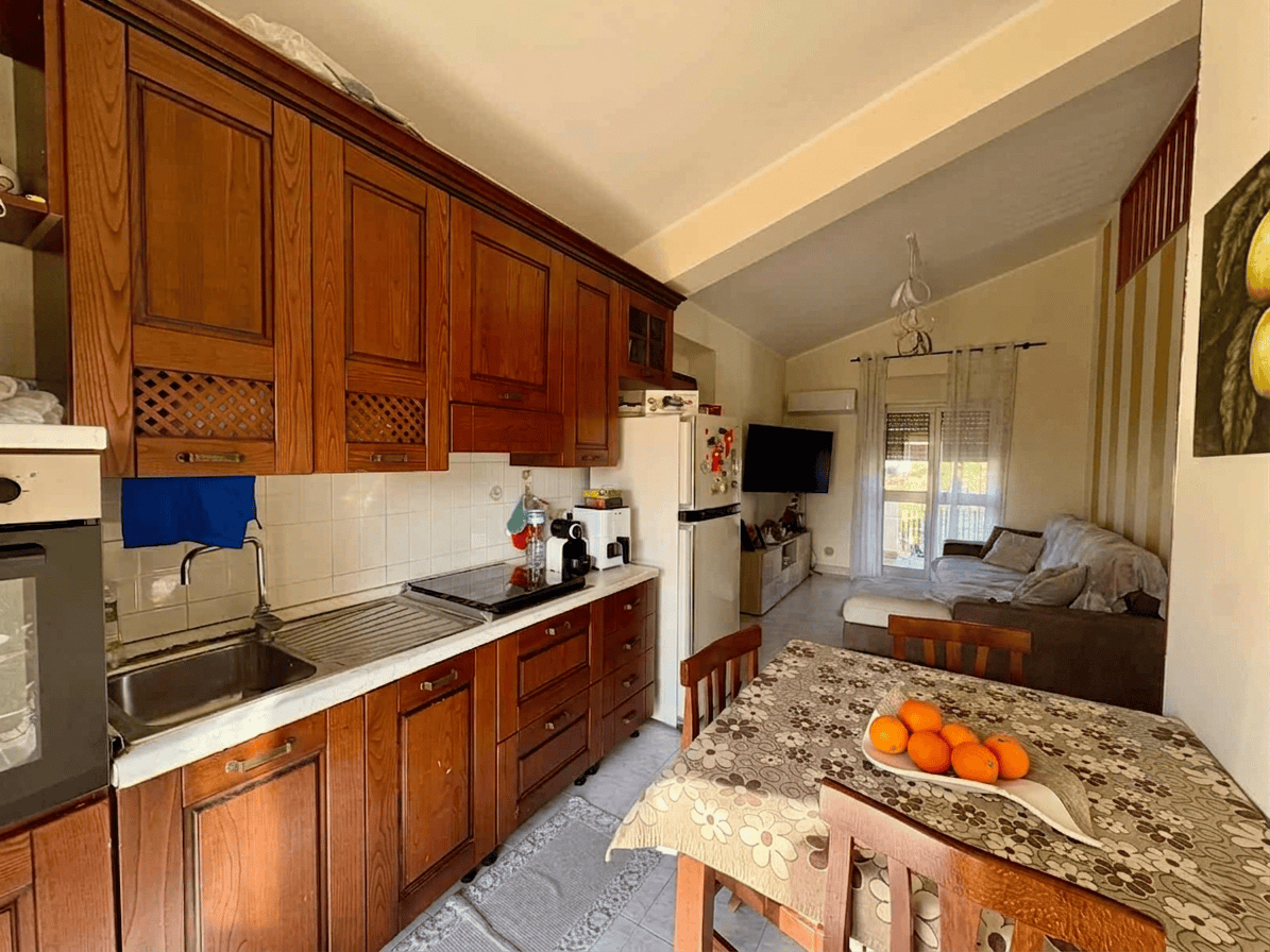 Cucina abitabile con mobili in legno, tavolo con tovaglia e frutta fresca, soggiorno con divano e TV, ambiente luminoso e accogliente, ideale per vivere e rilassarsi.