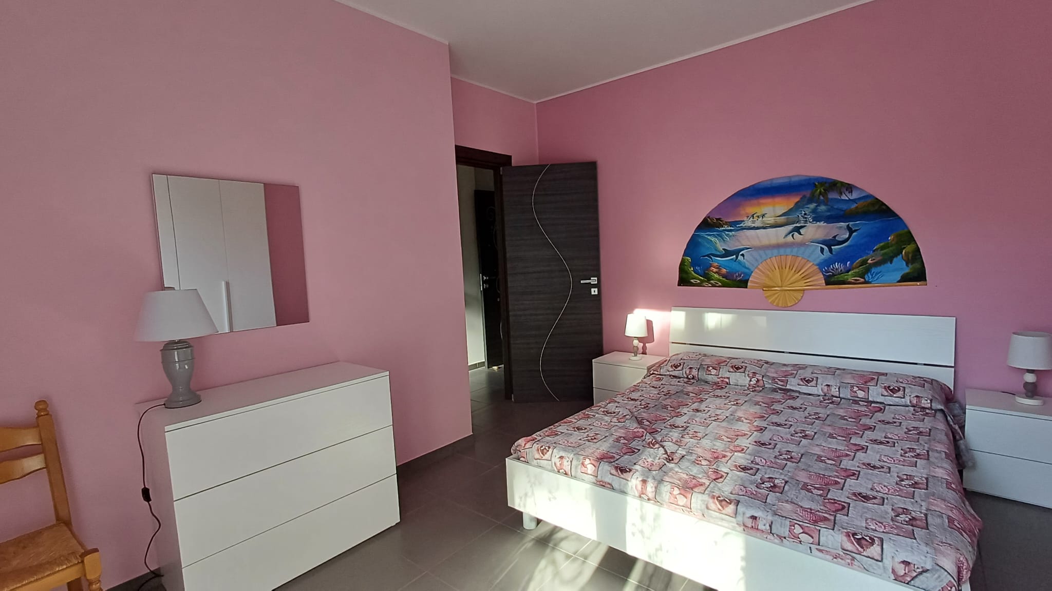 Foto di una camera da letto con pareti rosa, letto matrimoniale con copriletto decorato, lampade da tavolo, specchio e decorazione artistica di un fan con paesaggio marino.