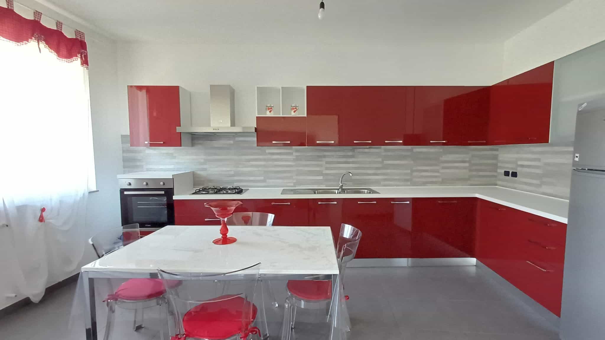 Cucina moderna con mobili rosso lucido, piano in vetro e elettrodomestici bianchi, ambiente luminoso con finestra e sedie trasparenti con seduta rossa. Ideale per living e attività quotidiane.