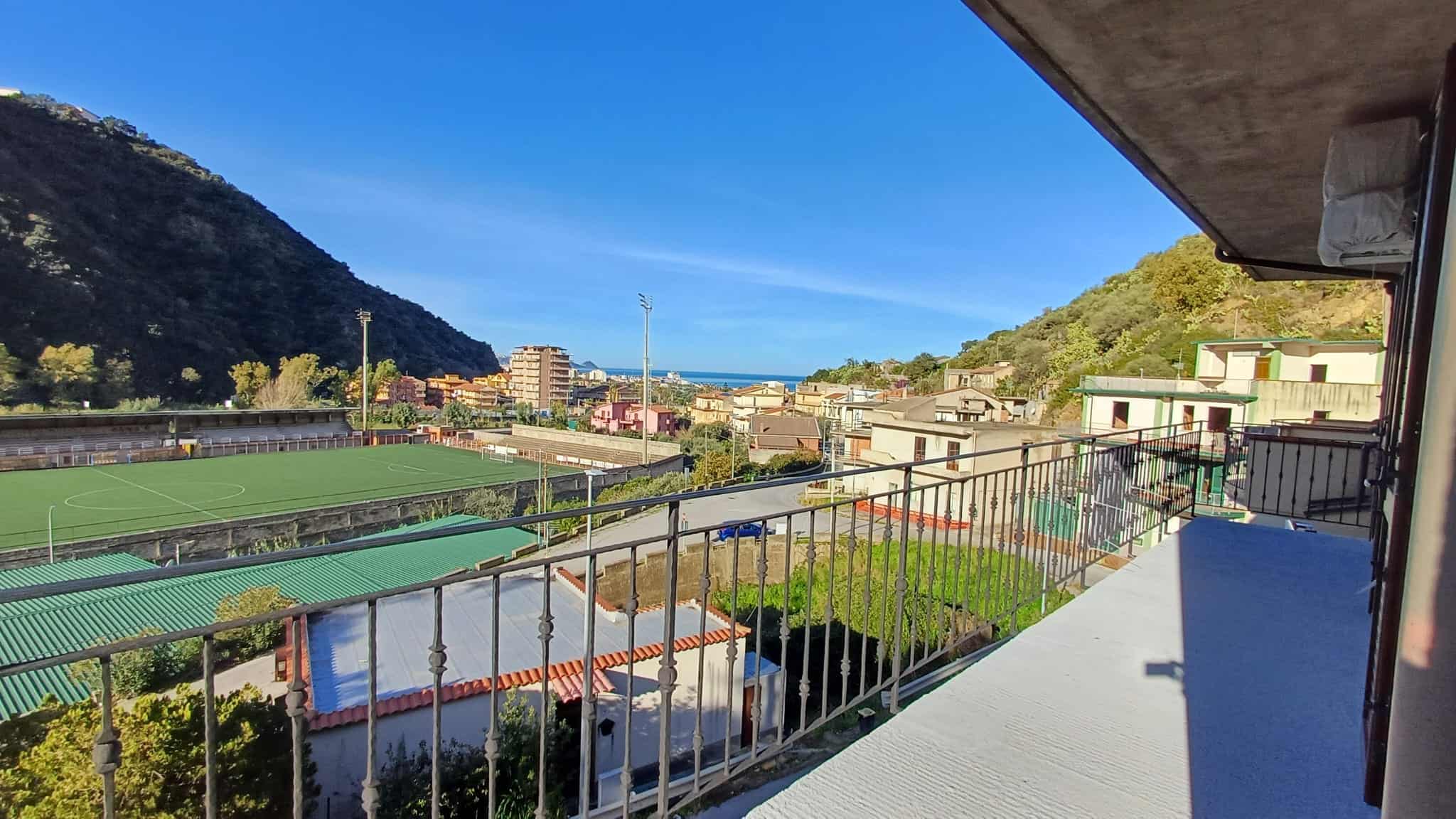 Vista panoramica sul campo da calcio, case colorate e montagne verdi in un appartamento con balcone a NextCasa, ideale per investimento immobiliare in zona tranquilla e ben servita.
