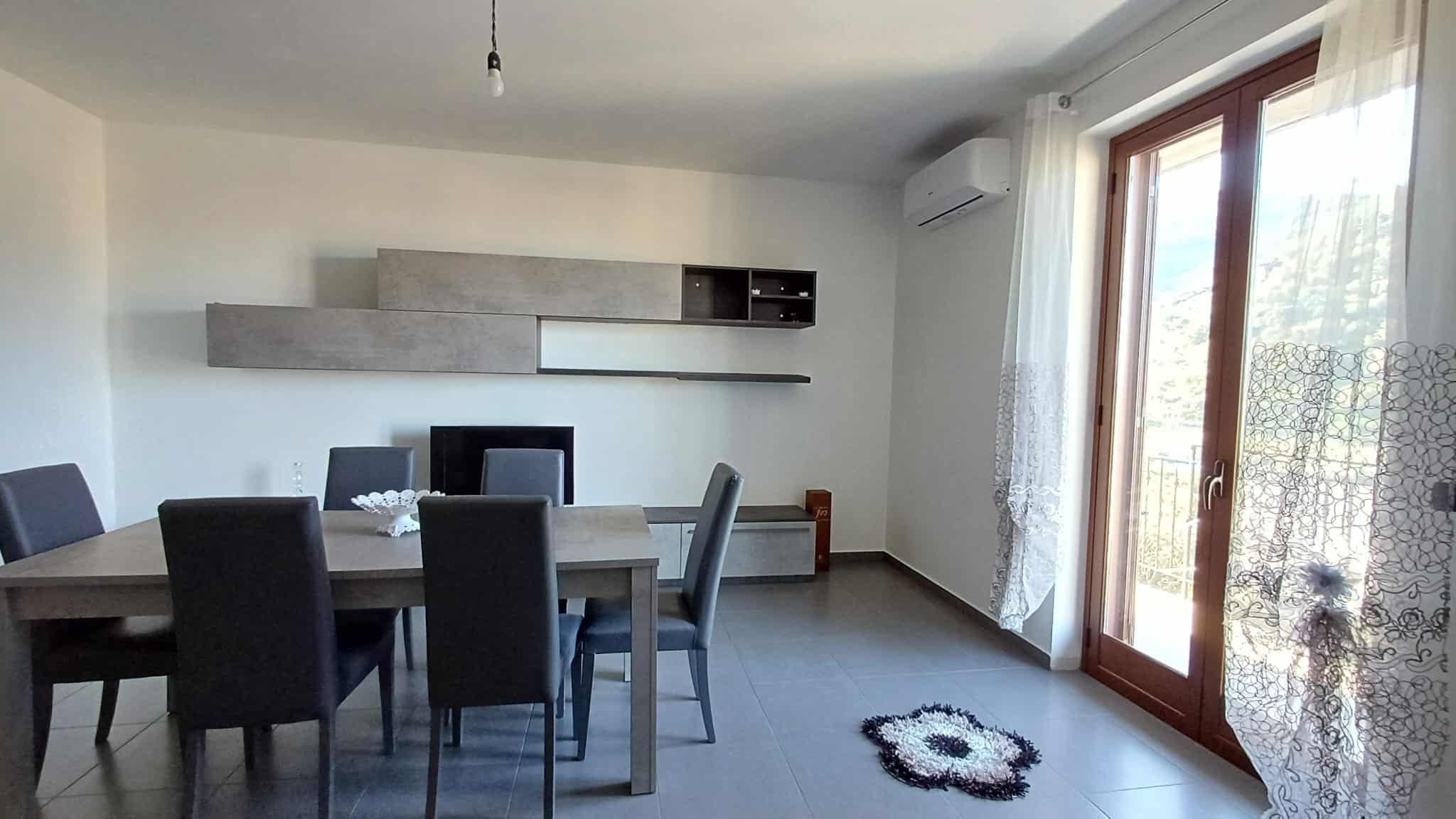 Vista di un soggiorno moderno con tavolo da pranzo, sedie in pelle grigia, parete attrezzata, grande finestra con tende e balcone, atmosfera luminosa, ideale per immobili di lusso e affitti residenziali.