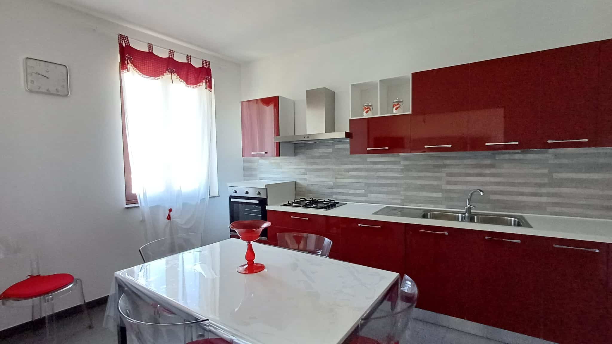 Cucina moderna in stile minimal con mobili rossi lucidi, piano cottura e lavabo in acciaio, ambientazione luminosa con finestra e tendina rossa, ideale per soluzioni abitative di design NextCasa.