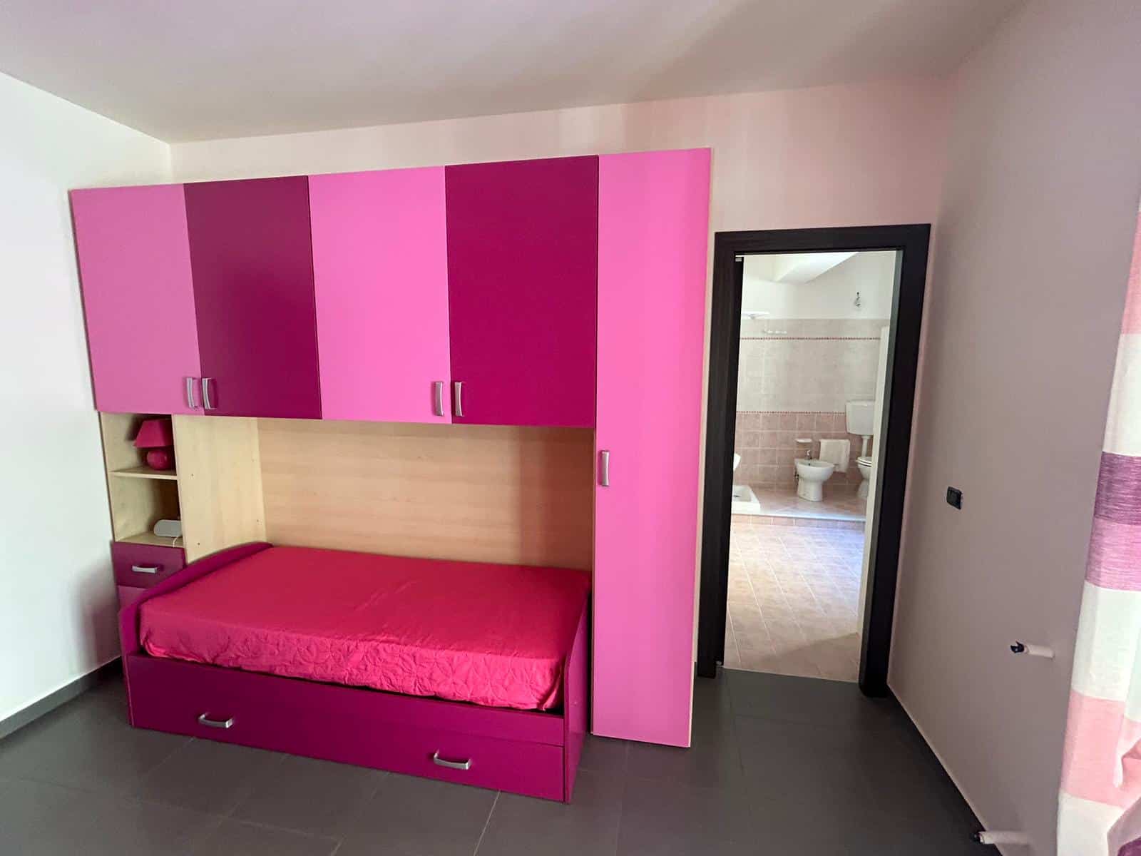 Letto a contenitore con armadio a muro rosa e bianco, vista dal soggiorno, camera da letto moderna e funzionale, ideale come soluzione hotel o affitto breve, in un'abitazione con bagno adiacente.