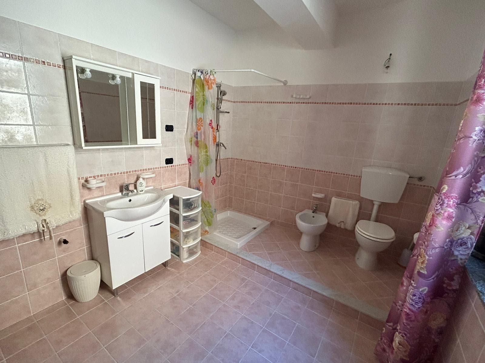 Bagno spazioso e luminoso con piastrelle rosa, lavabo, bidet, wc, doccia con tenda colorata, specchiera, arredi pratici e accessori funzionali, ideale per proprietà residenziali o investimento immobiliare.