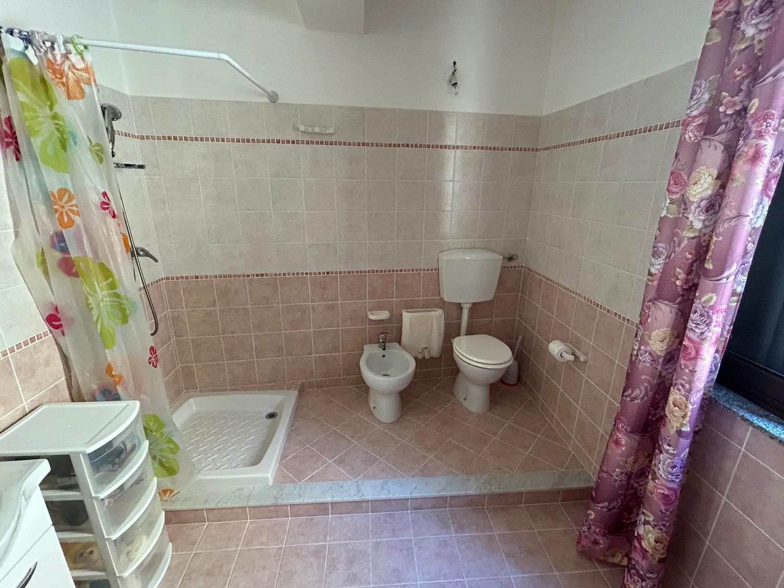 Lavabo, bidet e wc in un bagno con piastrelle rosa e accessori di arredamento normali, con finestra con tende floreali rosa, in un'immagine di NextCasa.