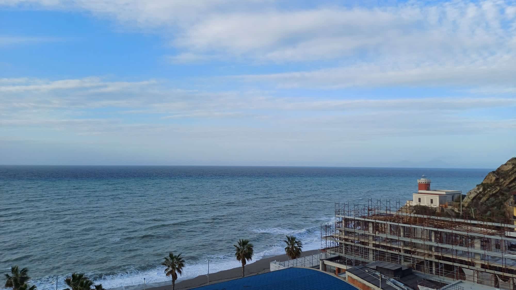 Vista panoramica mare con costa, costruzioni in fase di ristrutturazione, cielo sereno, palme sulla spiaggia, ideale per immobili fronte mare e proprietà di lusso a Napoli e Costiera Amalfitana.