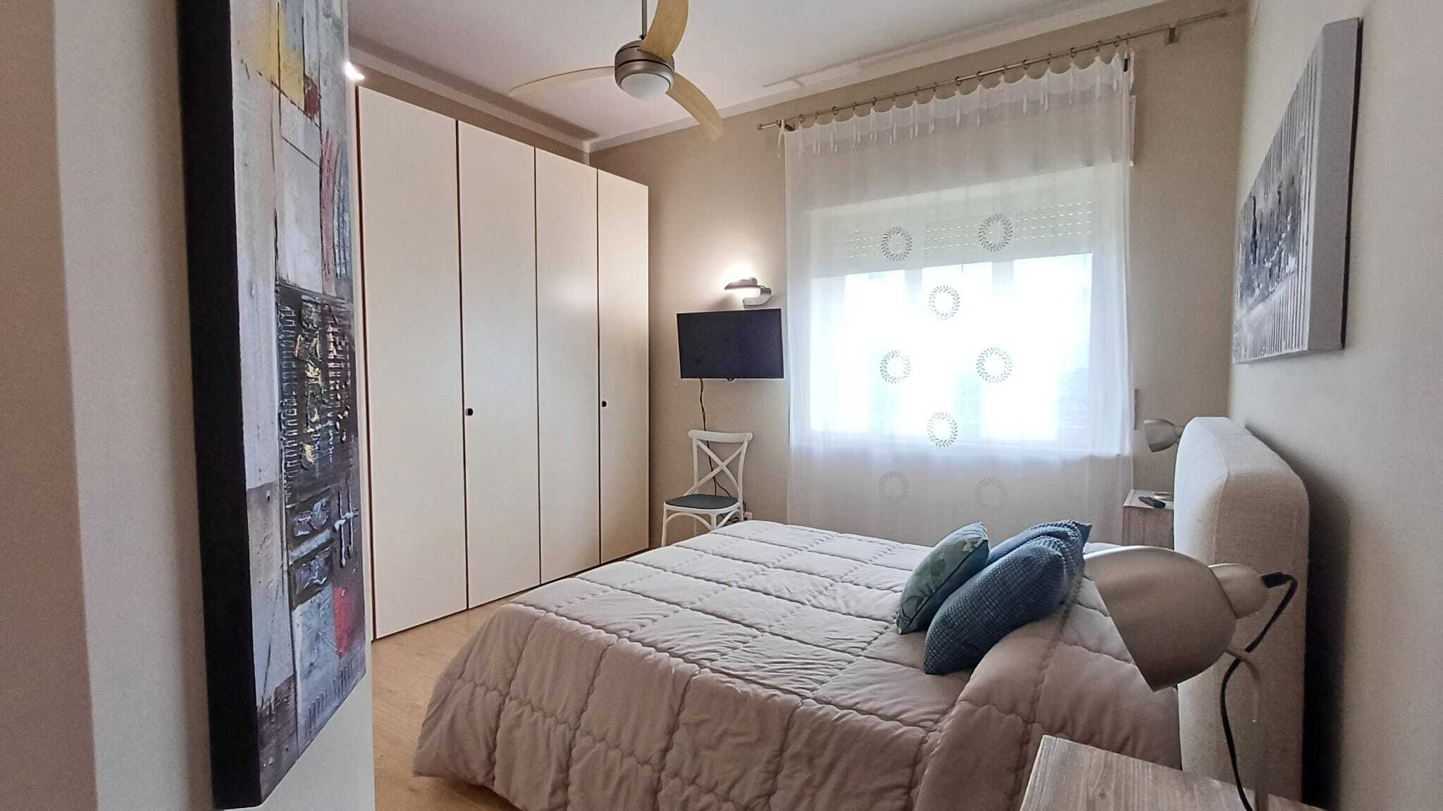 Riproduzione di una camera da letto moderna e accogliente con letto matrimoniale, armadio a muro, finestra con tende trasparenti e decorazioni artistiche, ideale per soluzioni abitative di qualità.