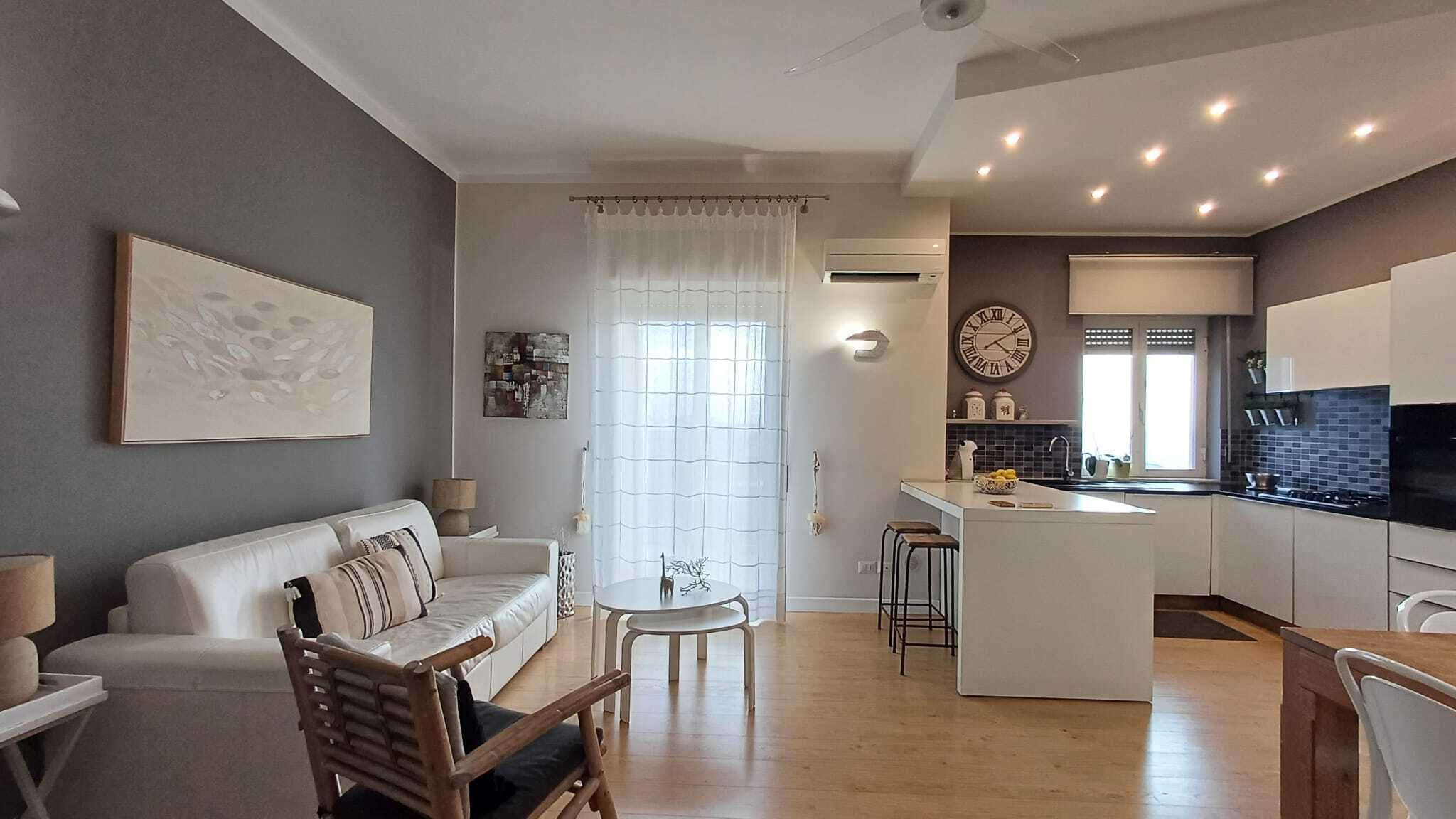 Appartamento moderno con soggiorno e cucina open space, pareti grigie e mobili bianchi, illuminazione a LED, decorazioni eleganti e ampie finestre con tende trasparenti, atmosfera accogliente e luminosa.