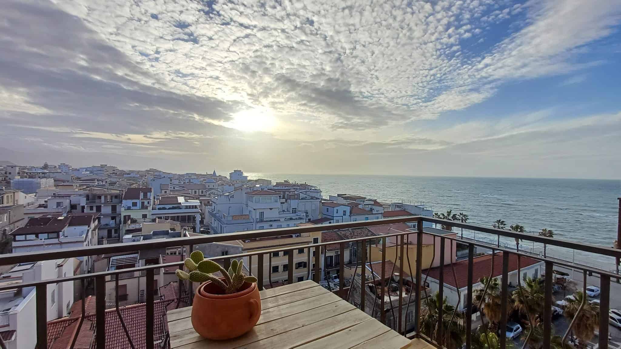 Vista panoramica sul mare e sulla città dal balcone di un appartamento vicino alla spiaggia, perfetto per case al mare e immobili in location marine, con un edificio in legno e pianta di cactus sul tavolo.