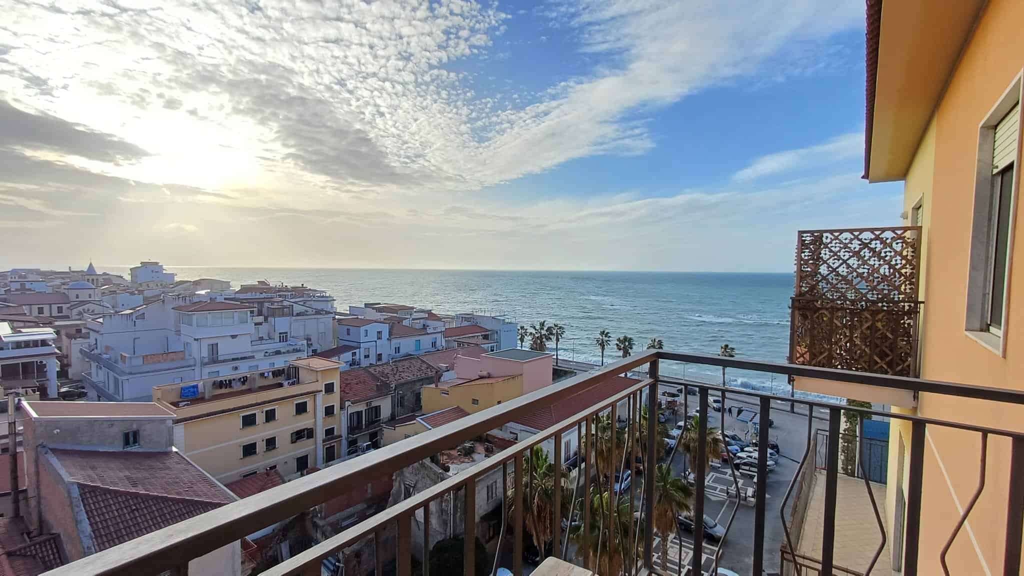 Vista mare dal balcone di un appartamento sul lungomare, con palme, case colorate e cielo nuvoloso, ideale per chi cerca immobili in vendita o affitto vicino al mare.