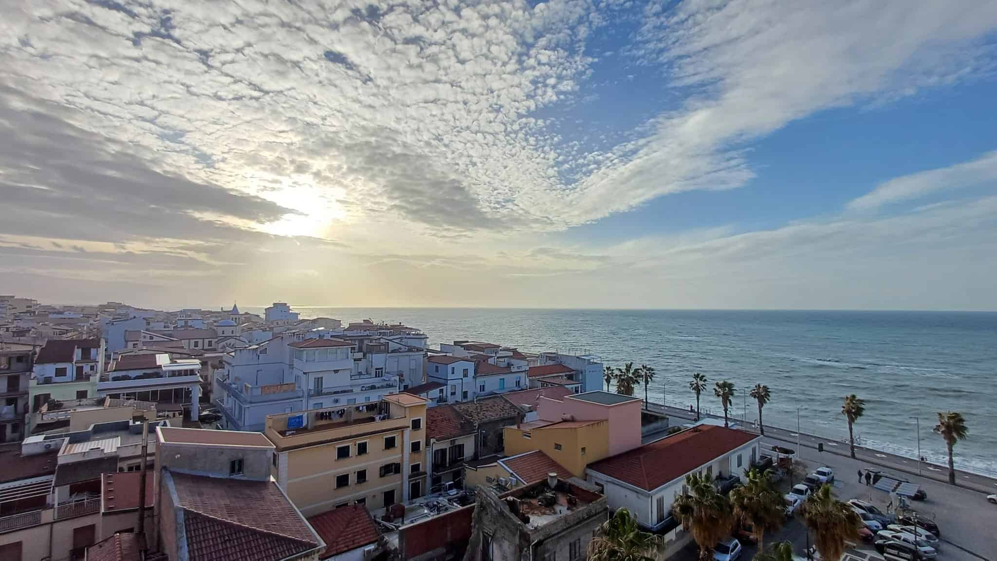 Vista mare con case sul lungomare e palme, al tramonto, a Napoli, Italia, vista dalla terrazza di un appartamento in vendita, con cielo nuvoloso e mare calmo, promuovendo l'acquisto di immobili fronte spiaggia.