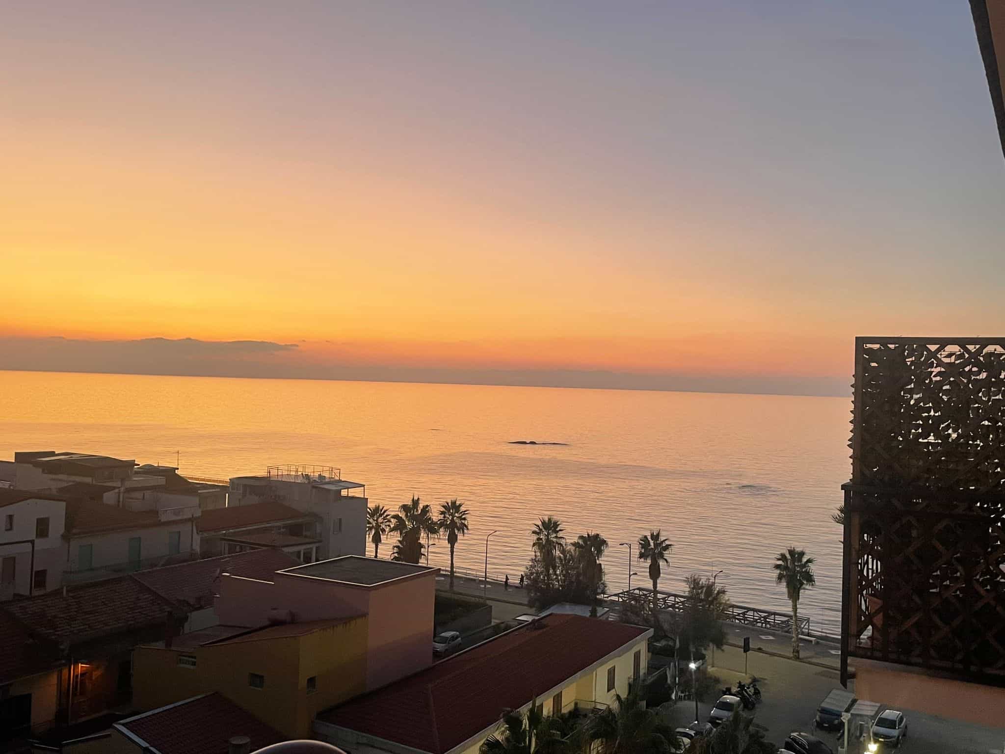 VISTA MARE AL TRAMONTO DA BALCONE DI APPARTAMENTO IN VENDITA IN PIEDI NELLA COSTA DELL'ITALIA, FOTOGRAFIA DI UNA CITTÀ COSTIERA CON PALME E EDIFICI COLORATI.