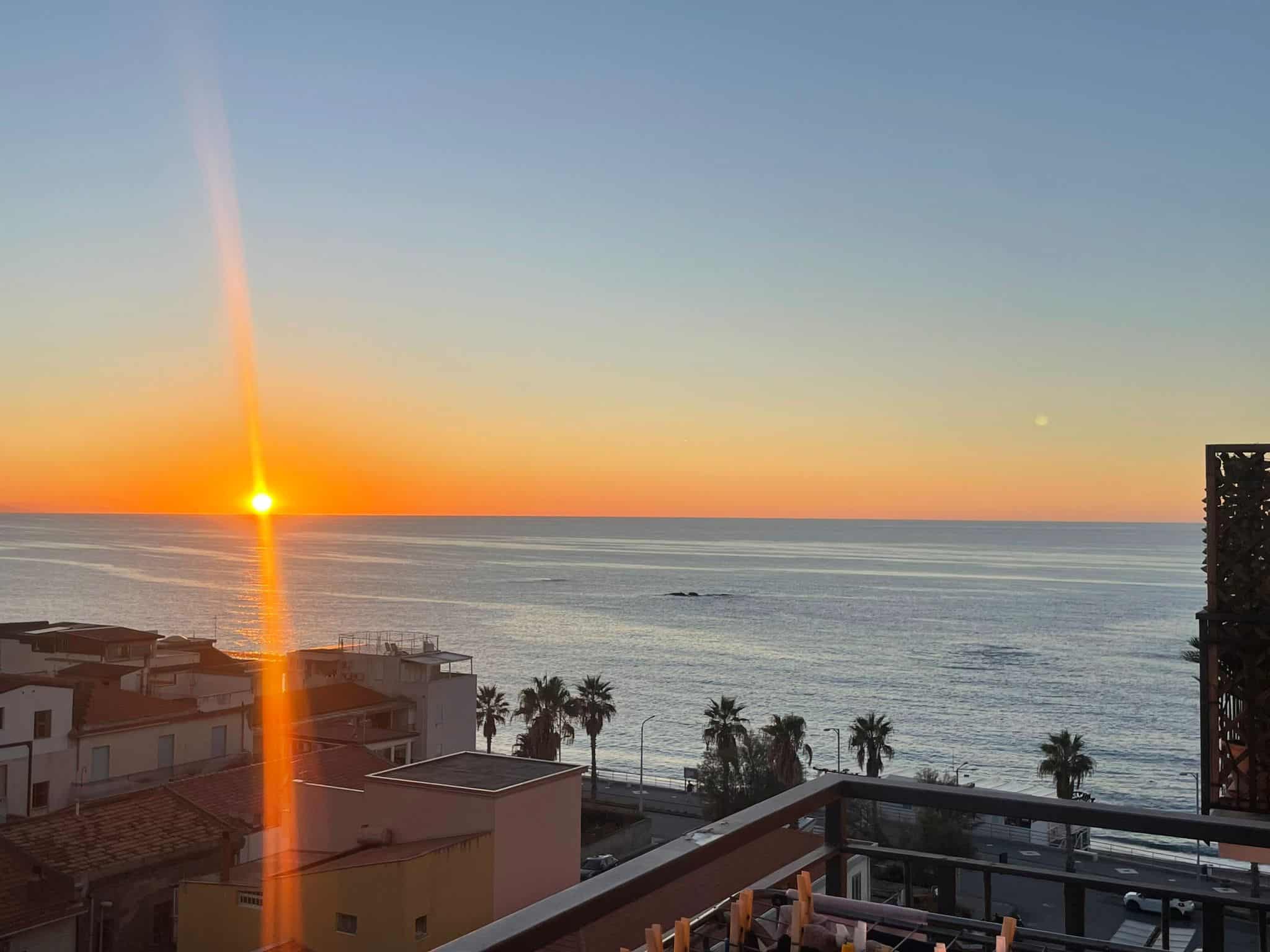 Vista mare al tramonto dalla terrazza di un appartamento a NextCasa, con palme e edifici residenziali, perfetta per immobili fronte mare e investimento immobiliare.