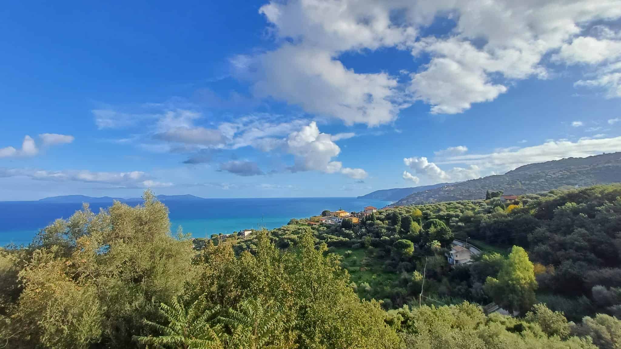 Vista panoramica dal mare con colline verdi, case e vegetazione mediterranea sotto un cielo azzurro con nuvole. Perfetta per immagini di immobili in vendita o affitto al mare.