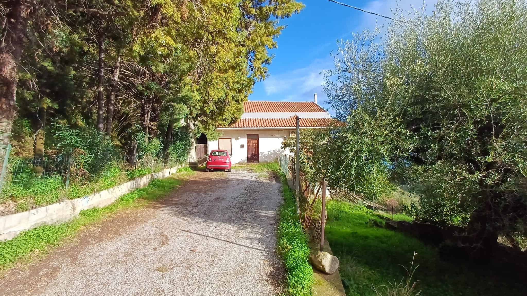 Auto in cortile di casa immersa nel verde con alberi e piante, strada sterrata, ambiente tranquillo, casa indipendente con porta in legno, tetto rosso e cielo sereno.