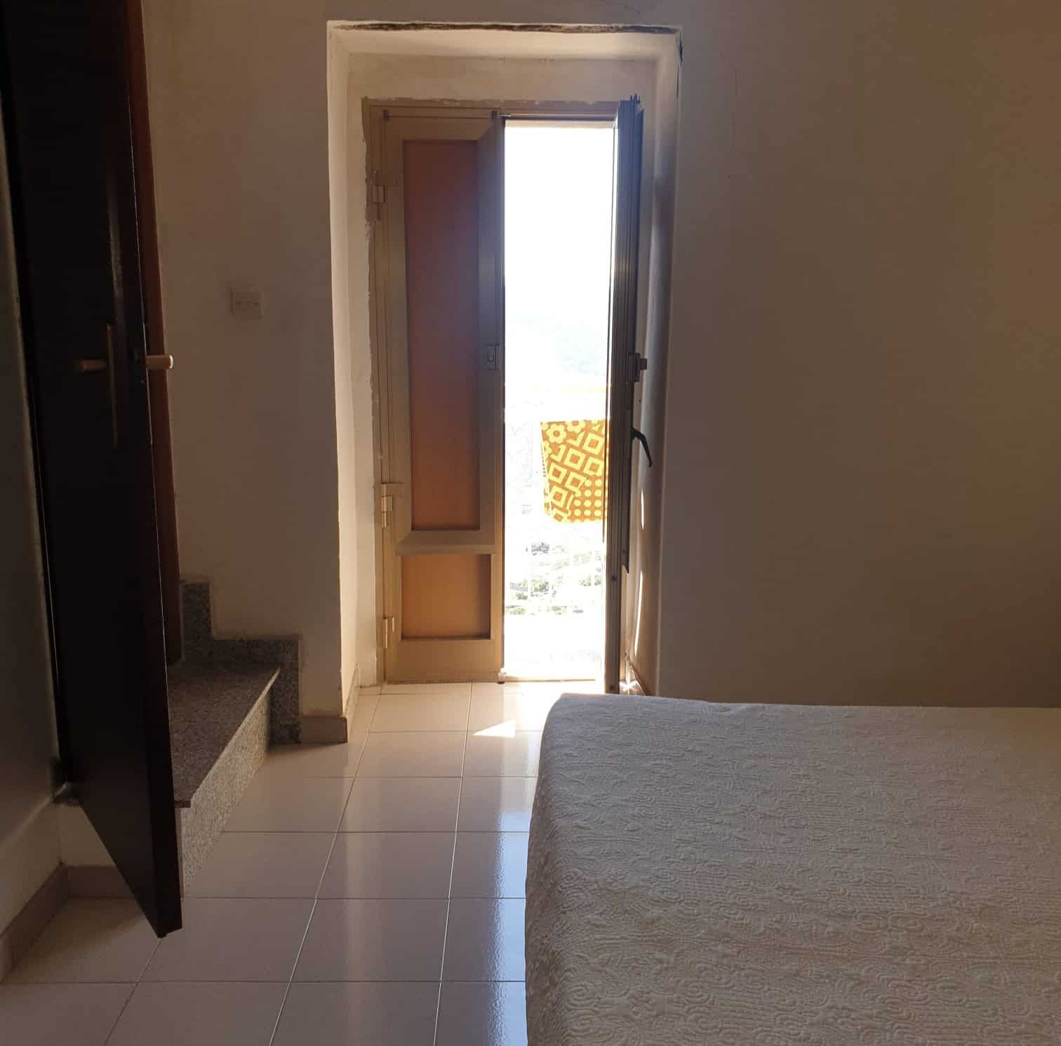Aria luminosa con porta finestra che conduce a un balcone, in una camera da letto moderna e accogliente, ideale per affitti turistici o residenziali, con pavimento in piastrelle chiare e pareti neutre.