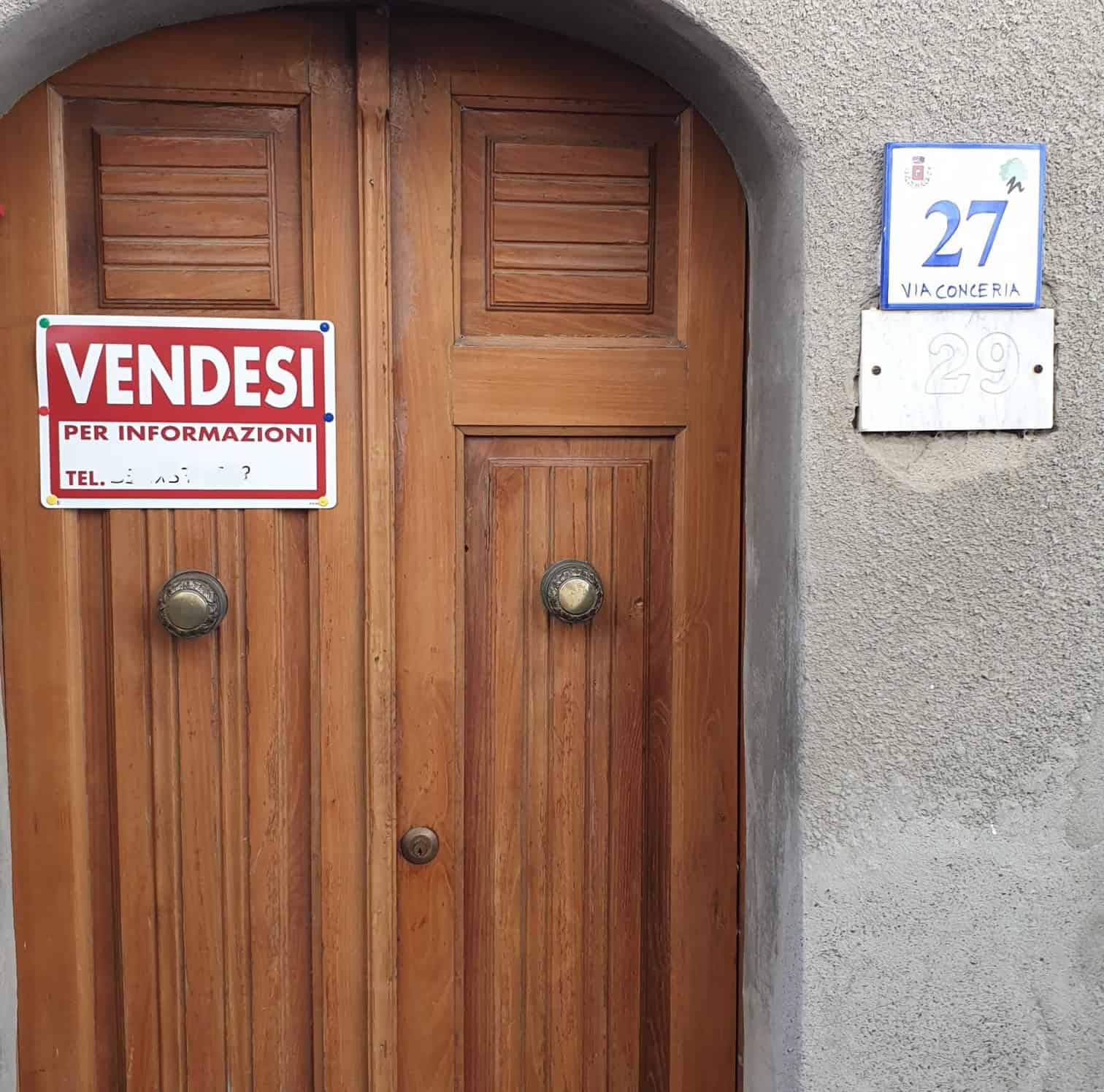 Porta in legno con cartello "VENDESI" e numeri civici 27 e 29, situata in una parete in cemento grigia, con dettagli di maniglie rotonde e un arco sopra l'ingresso, in un contesto urbano italiano.