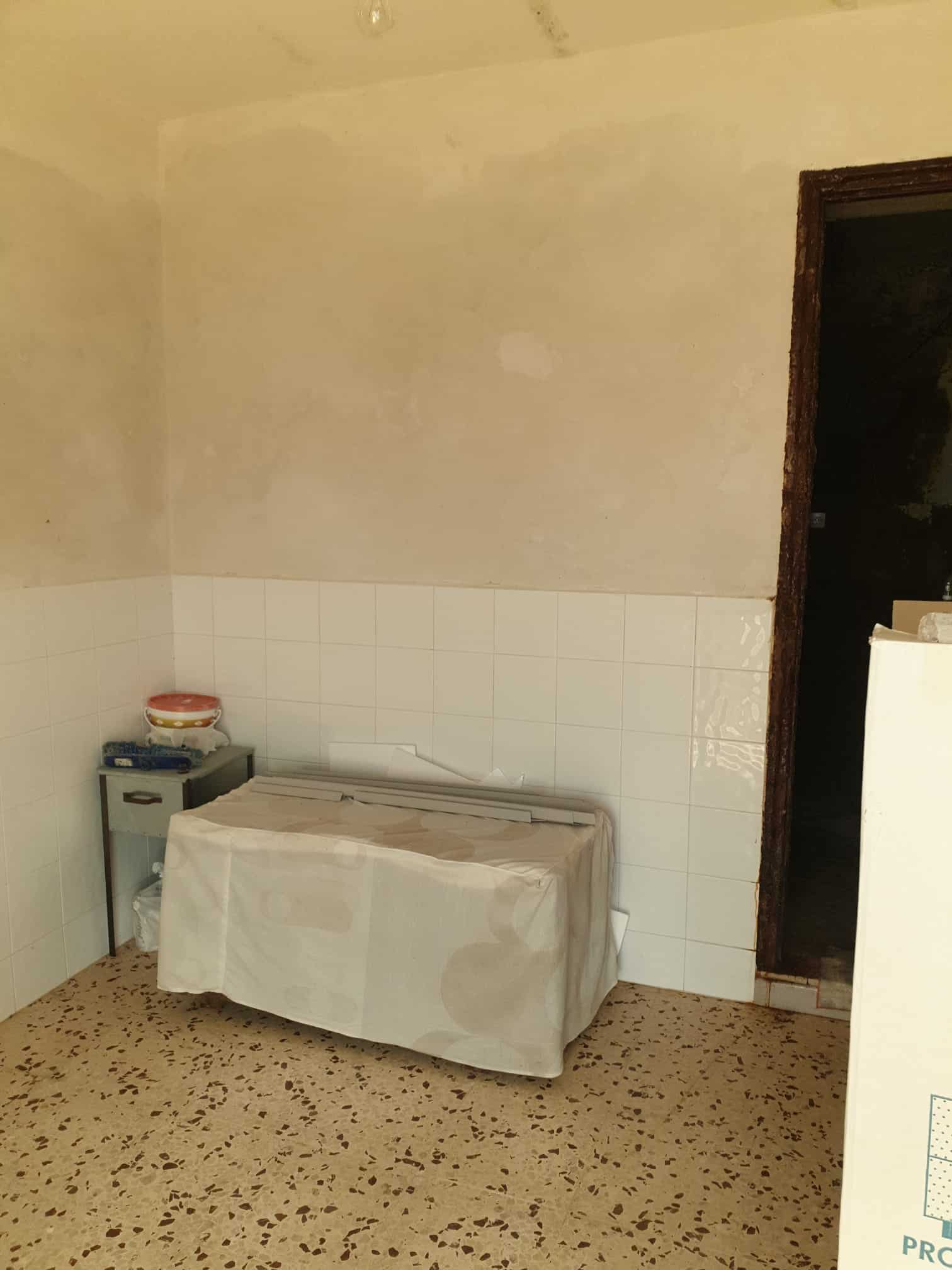 Spazio vuoto in una stanza con pareti beige, pavimento in graniglia e tavolo coperto con tovaglia bianca, vicino a un angolo con porta aperta, ambiente semplice e funzionale.
