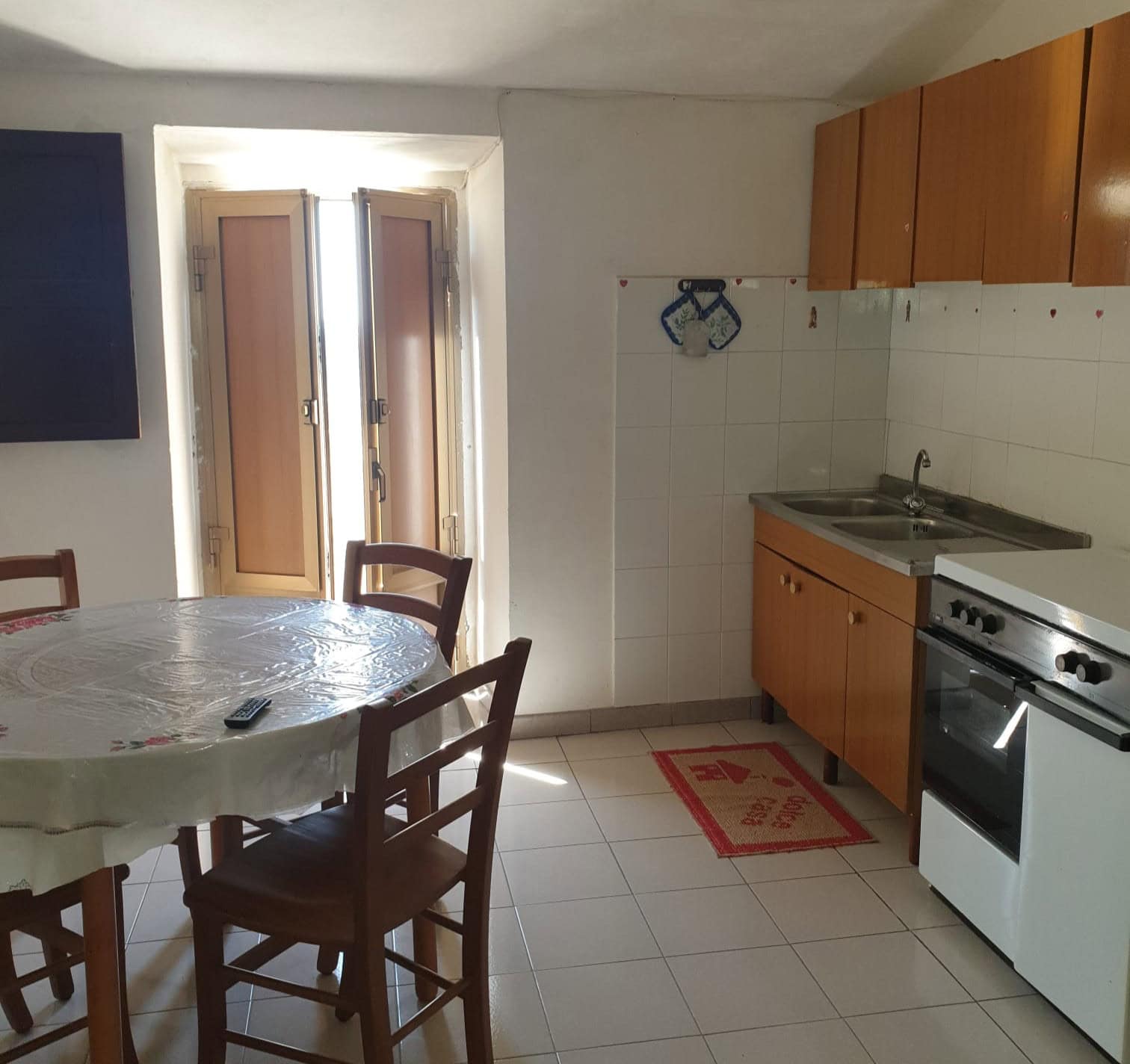 Cucina compatta con tavolo rotondo, sedie in legno, armadietti in legno chiaro, forno e lavabo, ambiente luminoso con finestra e porta finestra.