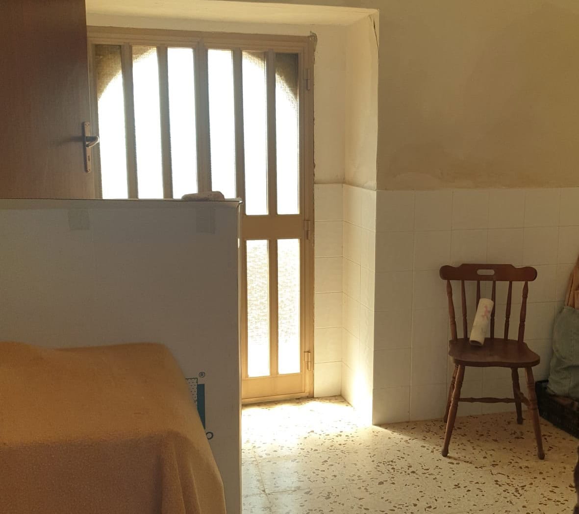 Interno di una stanza con porta finestra, sedia in legno e pareti in parte rivestite di piastrelle, ambiente luminoso e semplice.