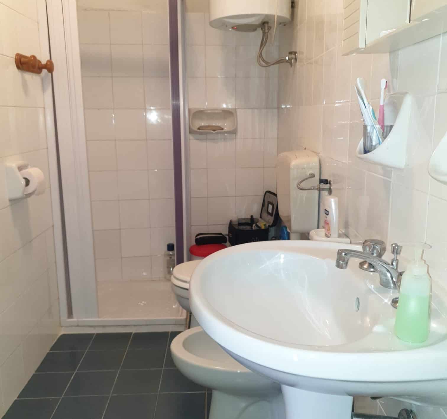 Lavabo e doccia in un bagno compatto con piastrelle chiare, accessori essenziali e spazio ottimizzato per l'uso quotidiano. Ideale per appartamenti moderni e funzionali.
