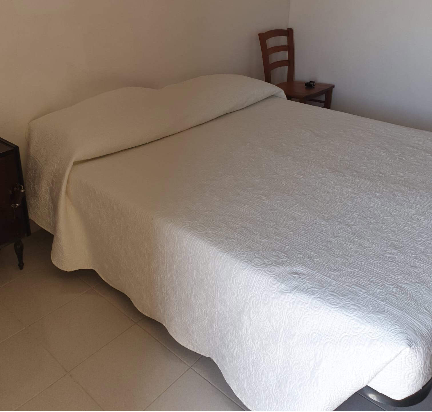 Lettino matrimoniale con copriletto bianco in camera da letto semplice e accogliente, con sedia in legno e pareti neutre, ideale per affitti turistici o residenziali.