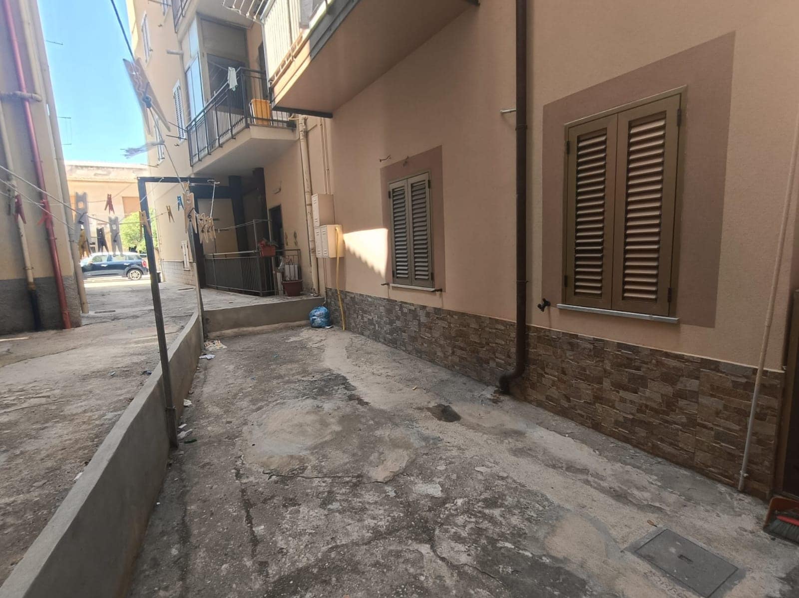 Riparazione e rinnovamento cortile condominiale con pavimentazione in cemento, facciata con rivestimento in pietra, finestre con persiane in legno, in contesto urbano adiacente a palazzine residenziali.