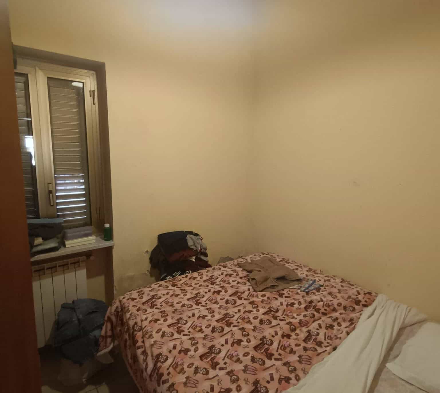 Camera da letto con finestra, letto con copriletto decorato, vestiti e oggetti vari sul letto, pareti beige, atmosfera semplice e informale.