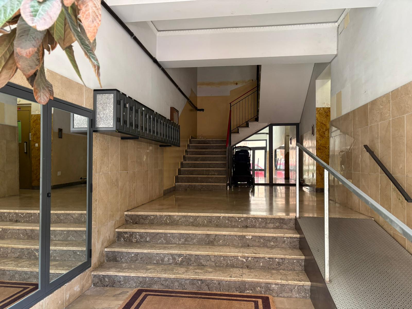 1. Ascensore e scalinate all'ingresso di un edificio residenziale, con mattonelle beige, rampella e porta con vetro, spazio accogliente e funzionale.
