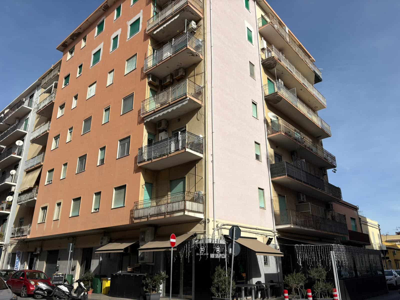 Vasto edificio residenziale con balconi e negozi al pian terreno, situato in zona urbana soleggiata, con parcheggio e attività commerciali vicine, ideale per immobili a disposizione di NextCasa.
