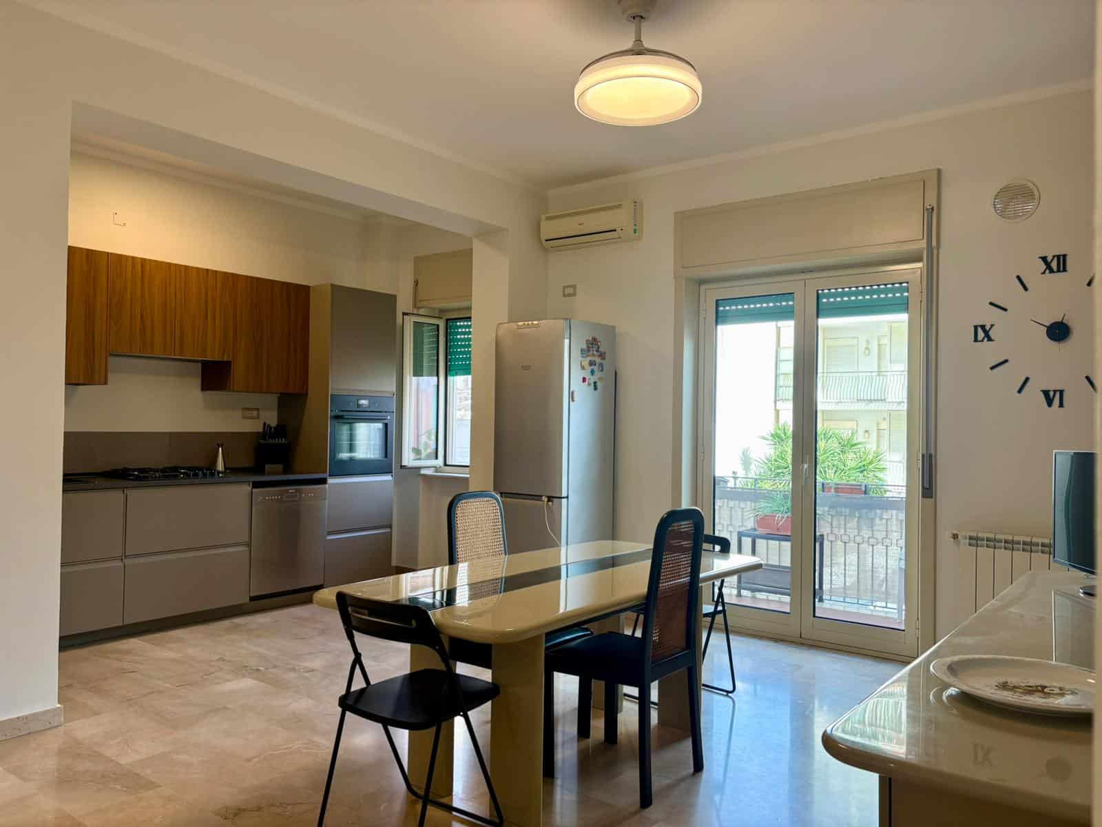 1. Cucina moderna con soggiorno aperto, mobili in legno e zona pranzo con tavolo e sedie, ampia finestra e balcone esterno.