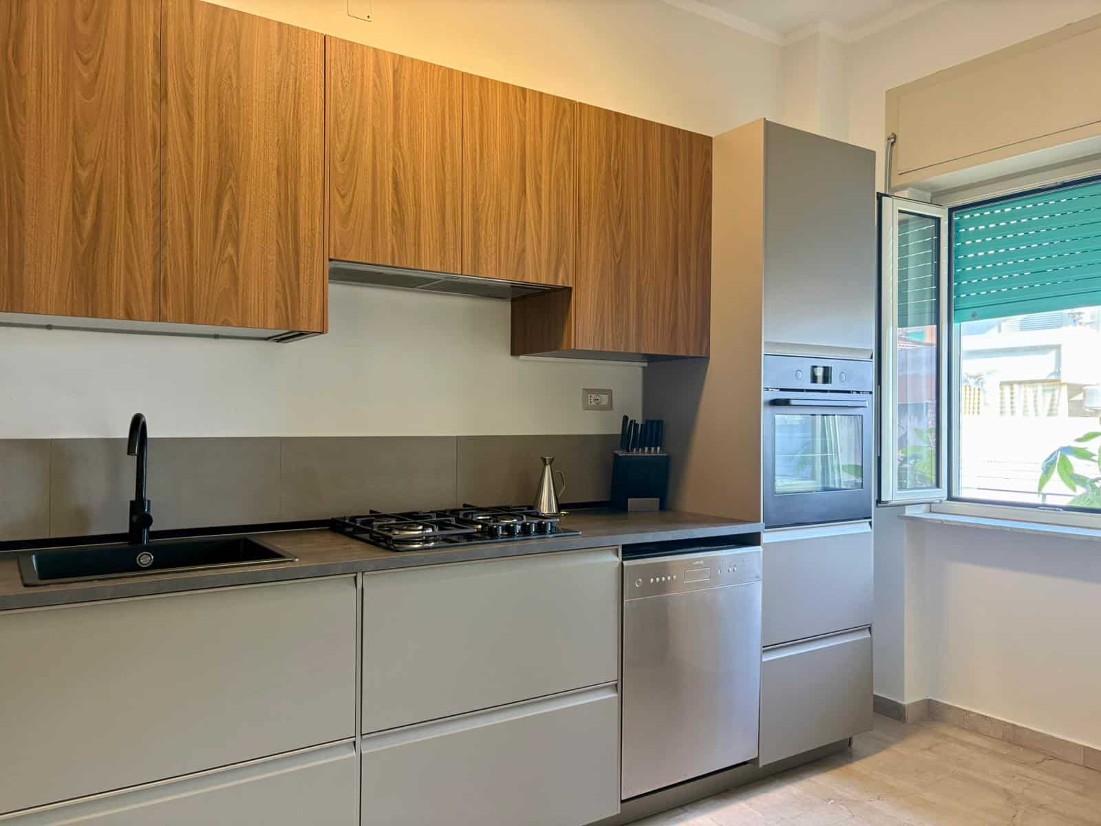 Cucina moderna con mobili in legno e elettrodomestici in acciaio, spaziosa e luminosa con finestra aperta verso l'esterno, ideale per appartamenti di design e comfort a NextCasa.