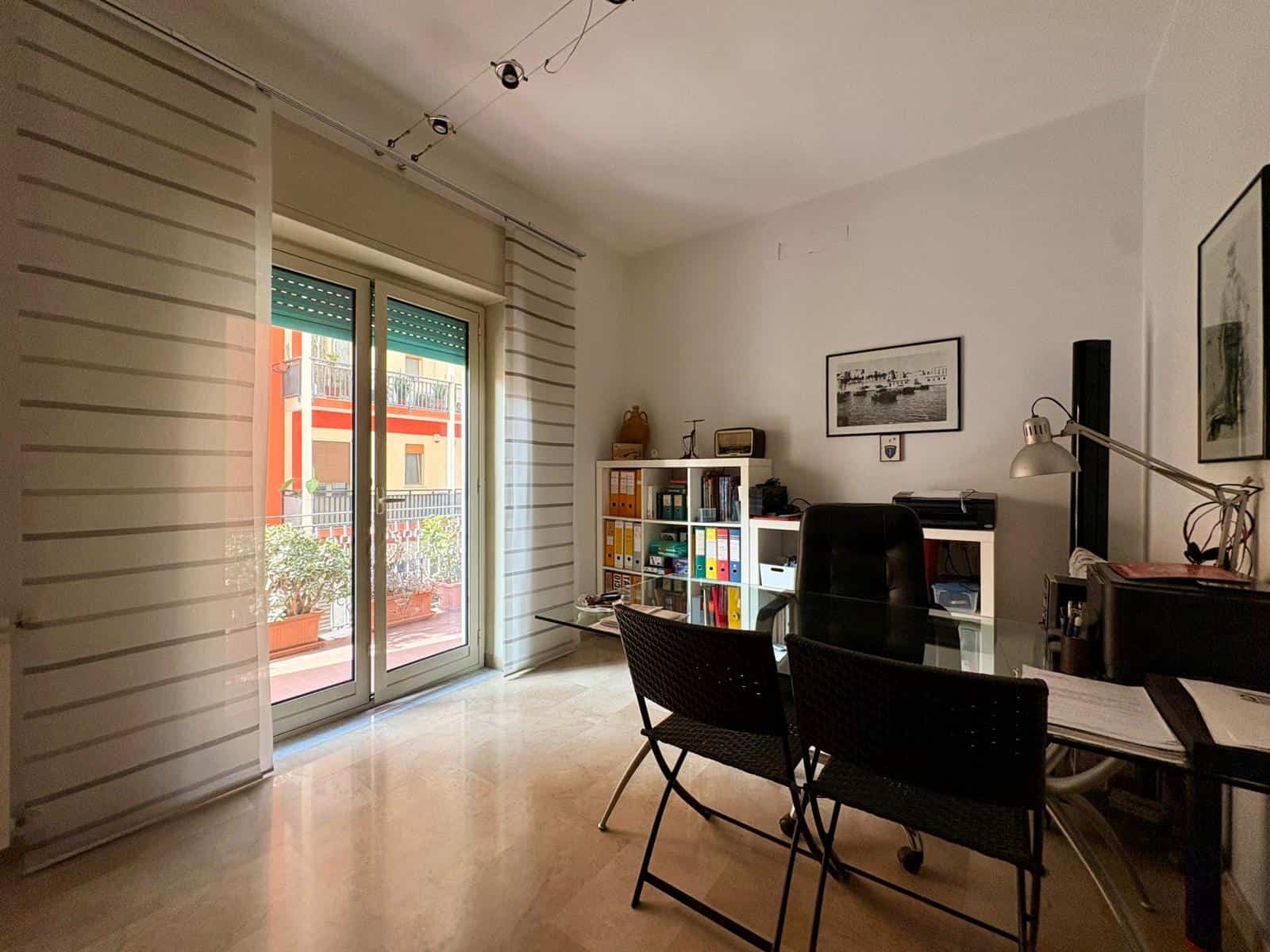 Stanza ufficio moderna con scrivania, sedia, scaffali, quadri e grande finestra che apre su balcone con piante, ambiente luminoso e funzionale per lavoro e studio.