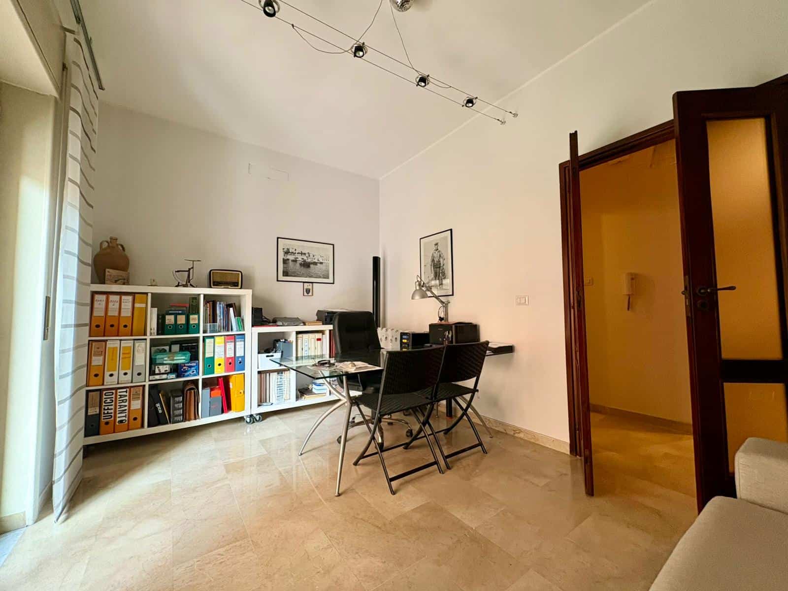 Spazio ufficio in casa con scrivania, sedie nere, libreria con cartelle colorate e oggetti decorativi, illuminato da luce naturale, ideale per lavoro e studio.