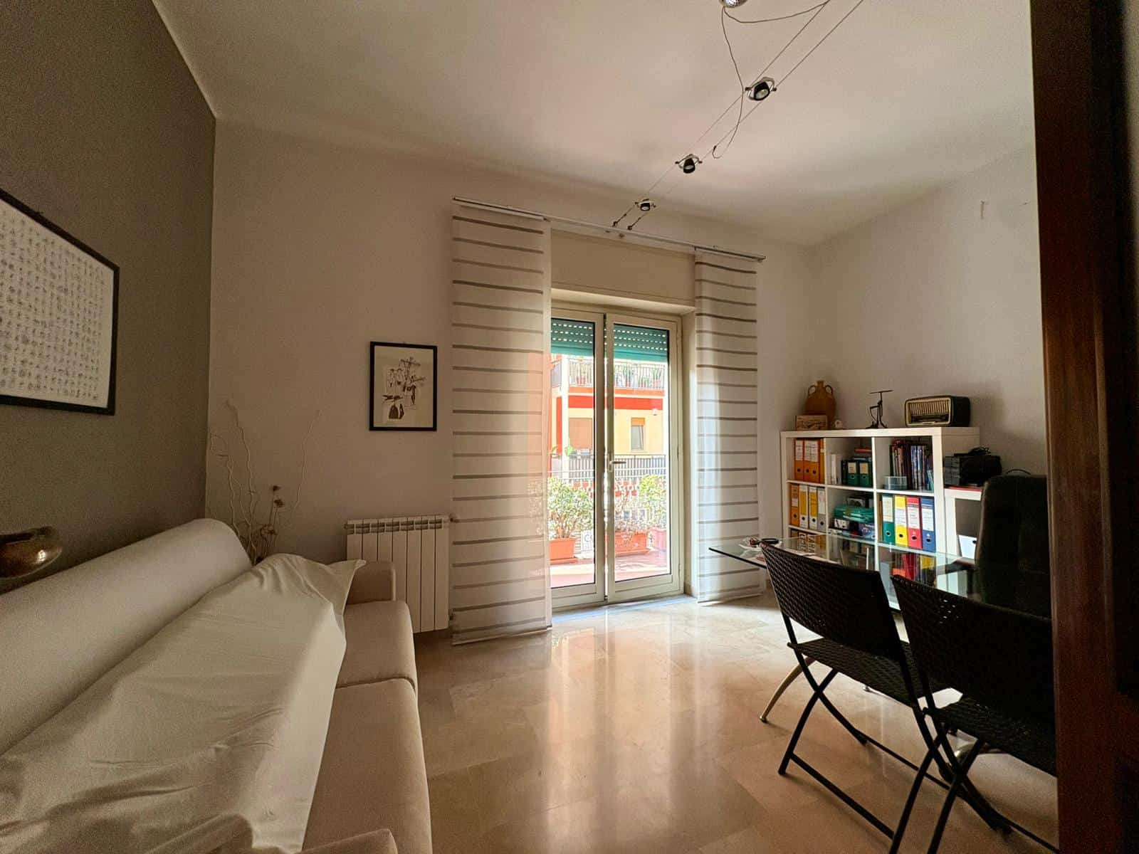 Immagine di soggiorno luminoso con divano elegante, vista su balcone con vista sulla città, arredamento funzionale con libreria colorata e illuminazione moderna, ideale per vivere in confort.