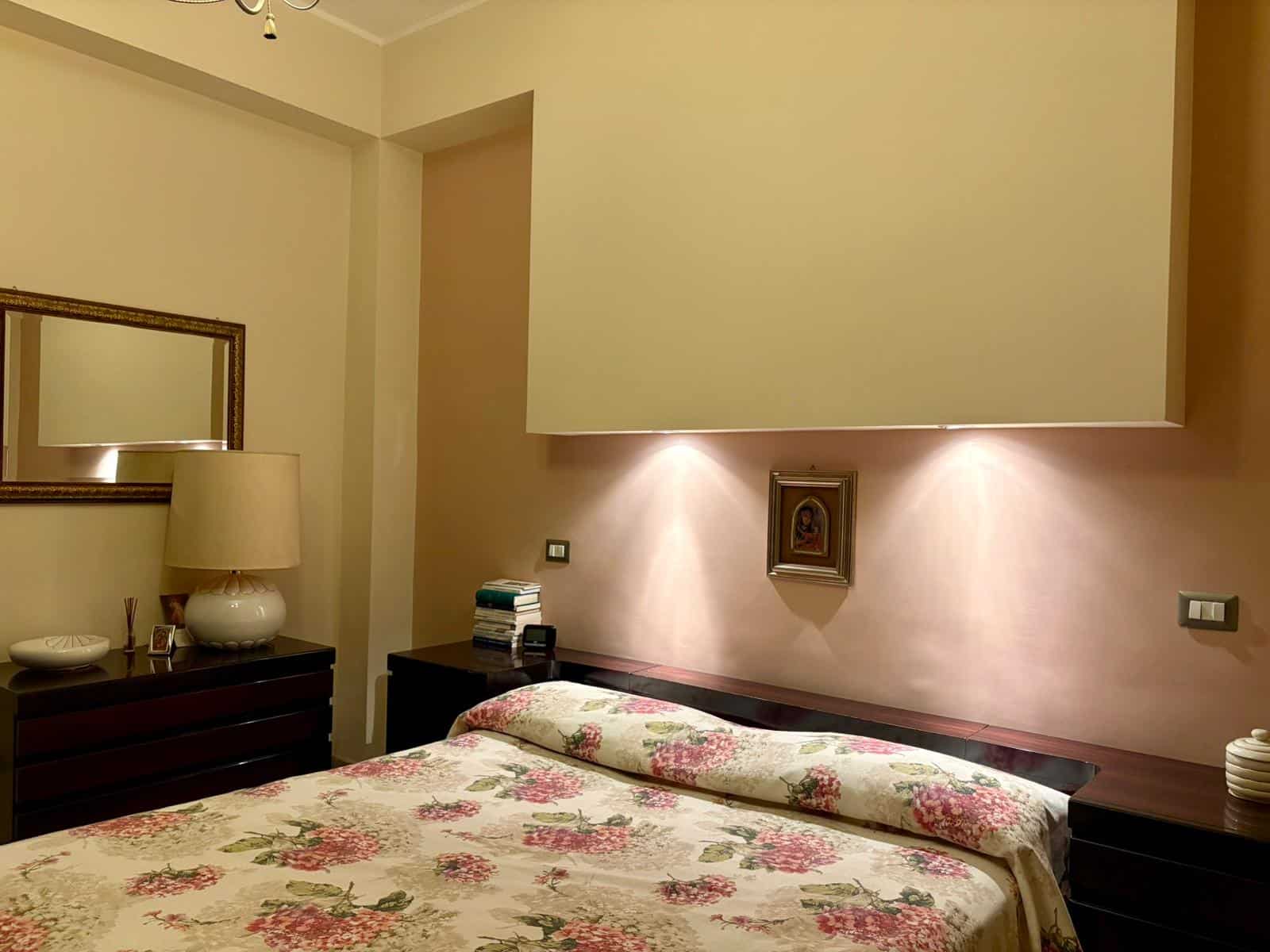 Finiture eleganti e atmosfera calda in camera da letto moderna con pareti beige, letti con trapunta floreale, illuminazione soffusa e arredi di design, ideale per servizi immobiliari e case di lusso.