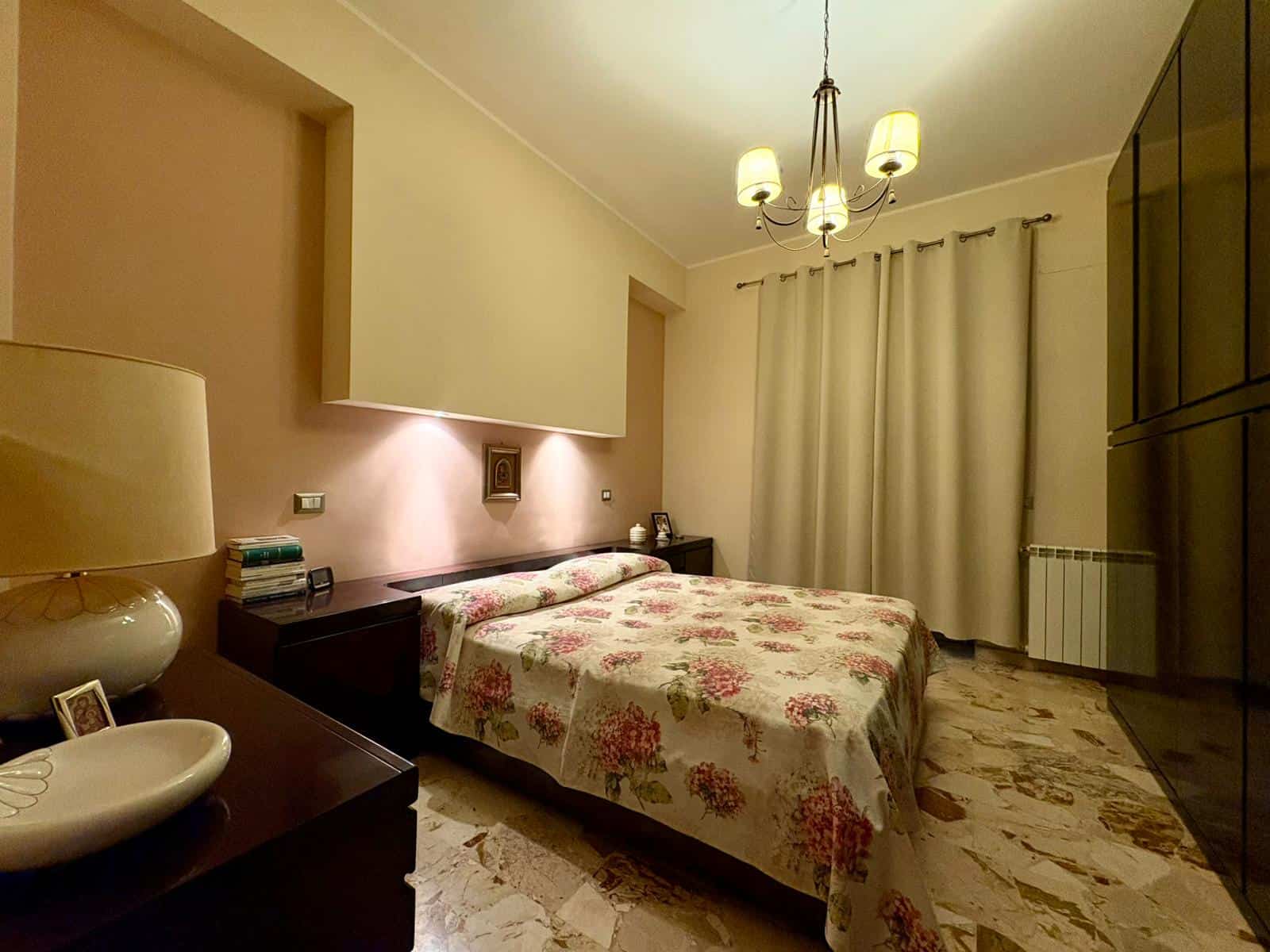 Fronte camera da letto moderna con letto matrimoniale, pareti color crema, lampadario elegante e armadio a muro, ambiente accogliente e ben illuminato per la casa dei sogni.