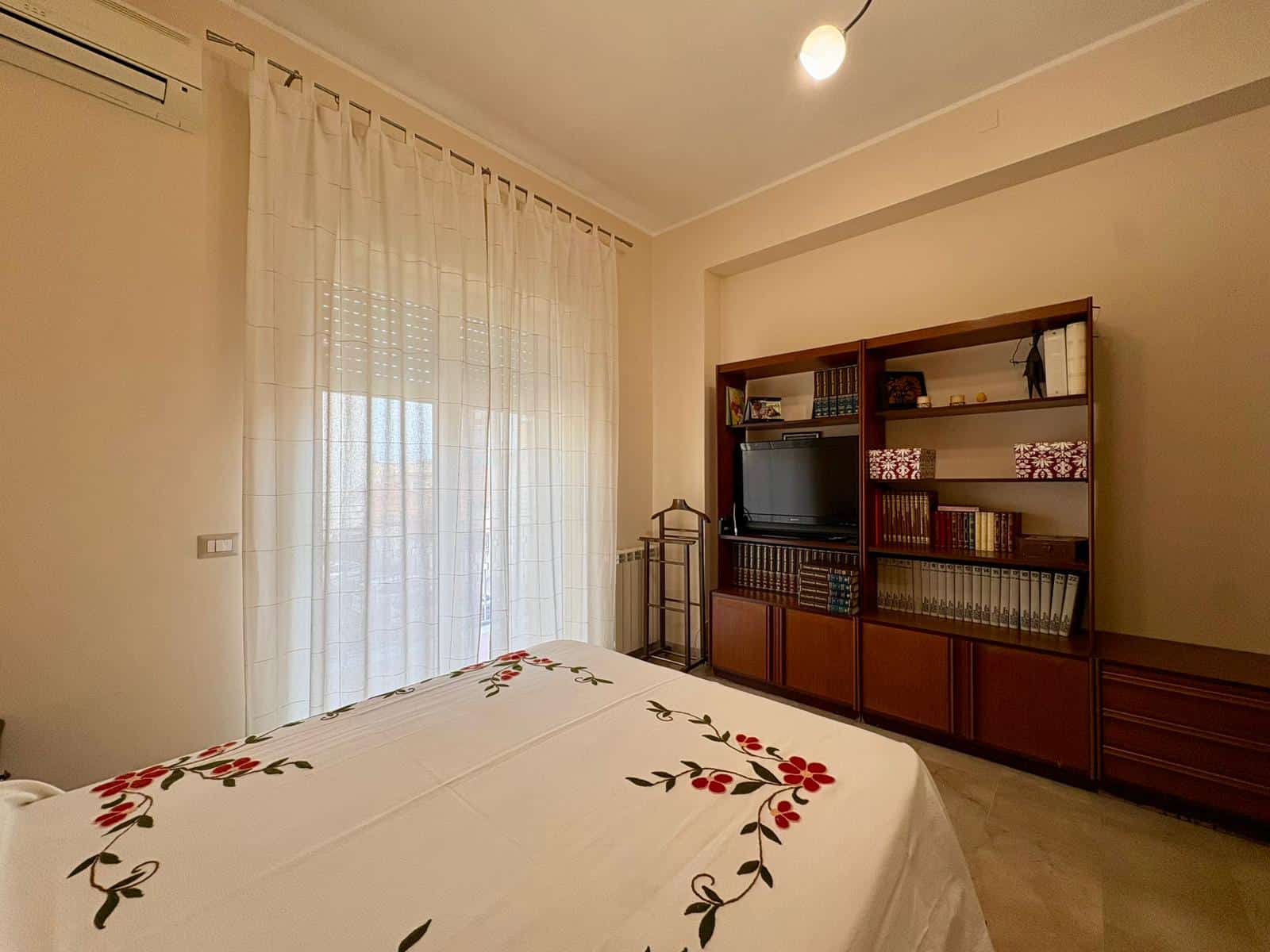 Letto matrimoniale con copriletto ricamato, soggiorno con mobiletto porta TV e libreria in legno, luce naturale da finestre con tende bianche, ambienti luminosi e accoglienti.