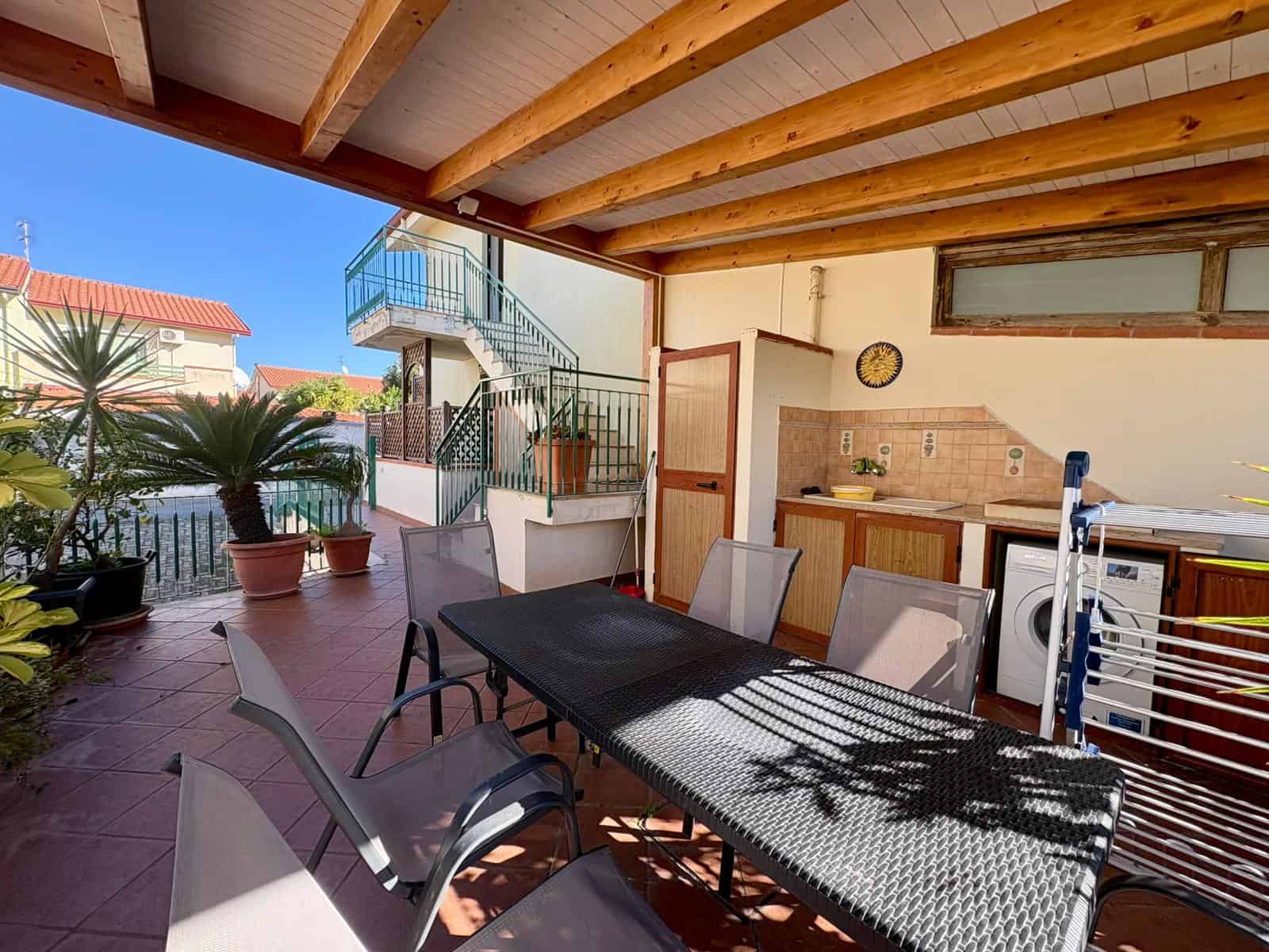 Spazio esterno coperto con tavolo e sedie, zona barbecue, piante in vaso e vista sulle case circostanti, ideale per momenti di relax e convivialità all'aperto.