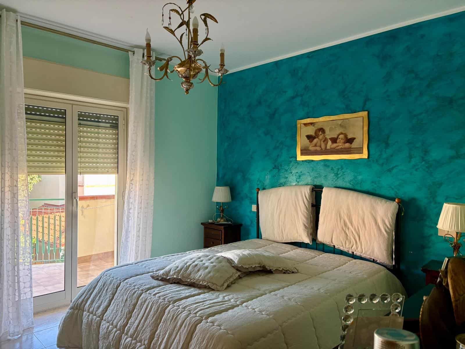 1. Colore pareti camera da letto turchese, arredamento classico e lampadario elegante, vista sul balcone con tende trasparenti, ambiente accogliente e luminoso di stile mediterraneo.