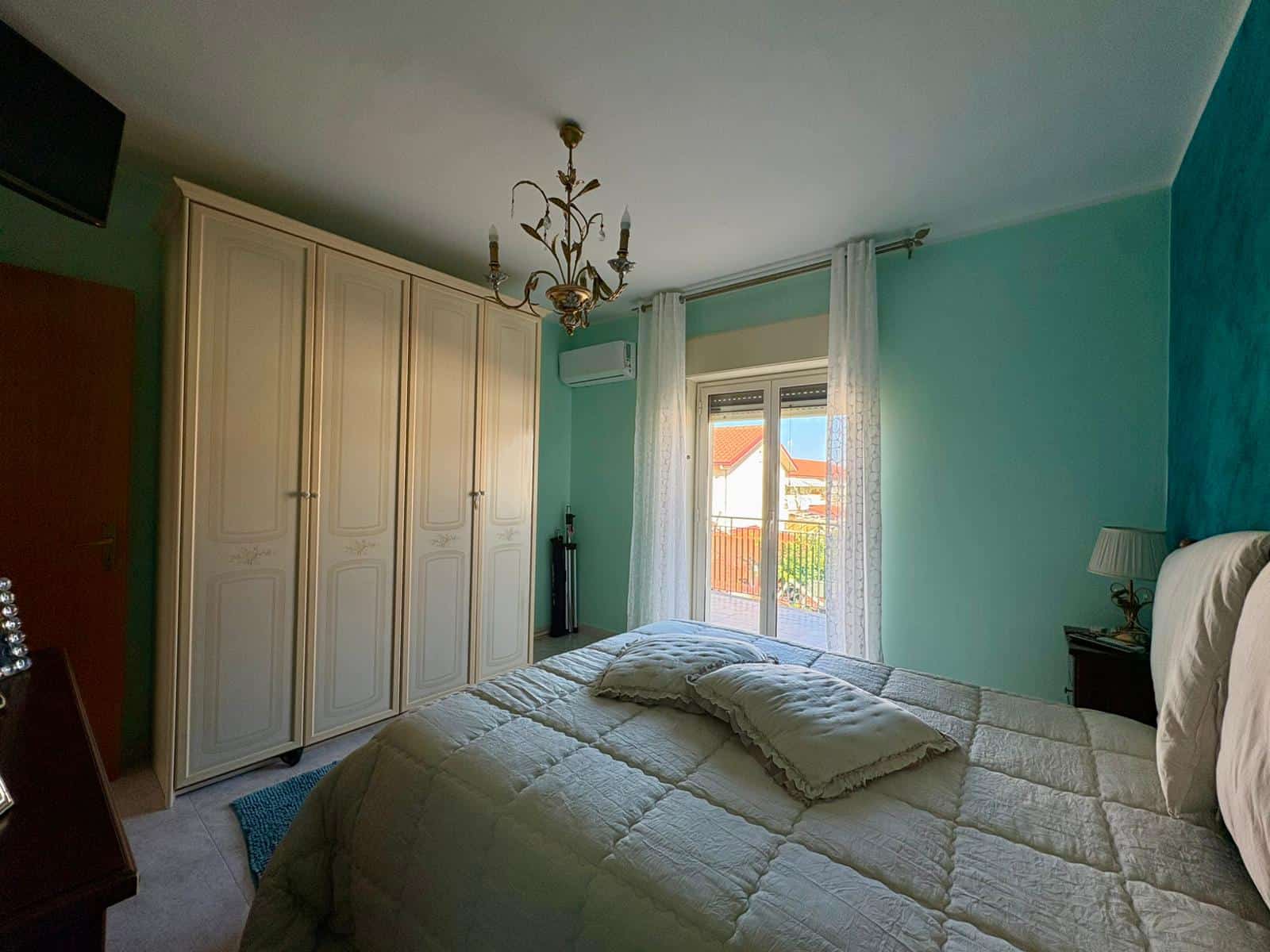 Ampia camera da letto con armadio a sei ante e balcone, arredamento classico, pareti verde acqua, lampadario elegante, lampada da tavolo, vista luminosa sul balcone con case e tetti rossi.