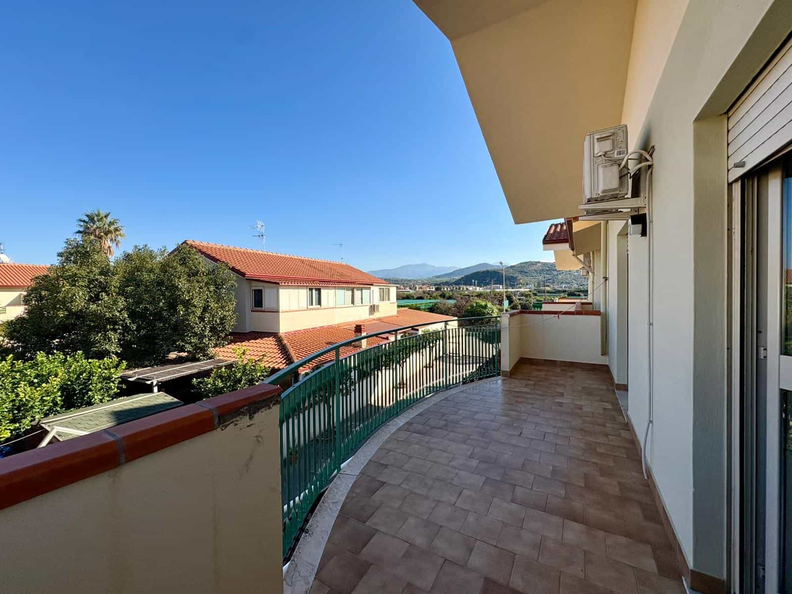 Vista panoramica di un ampio balcone di appartamento con vista su case con tetti rossi e montagne sullo sfondo, ambiente soleggiato e perfetto per relax e godersi l'estate.
