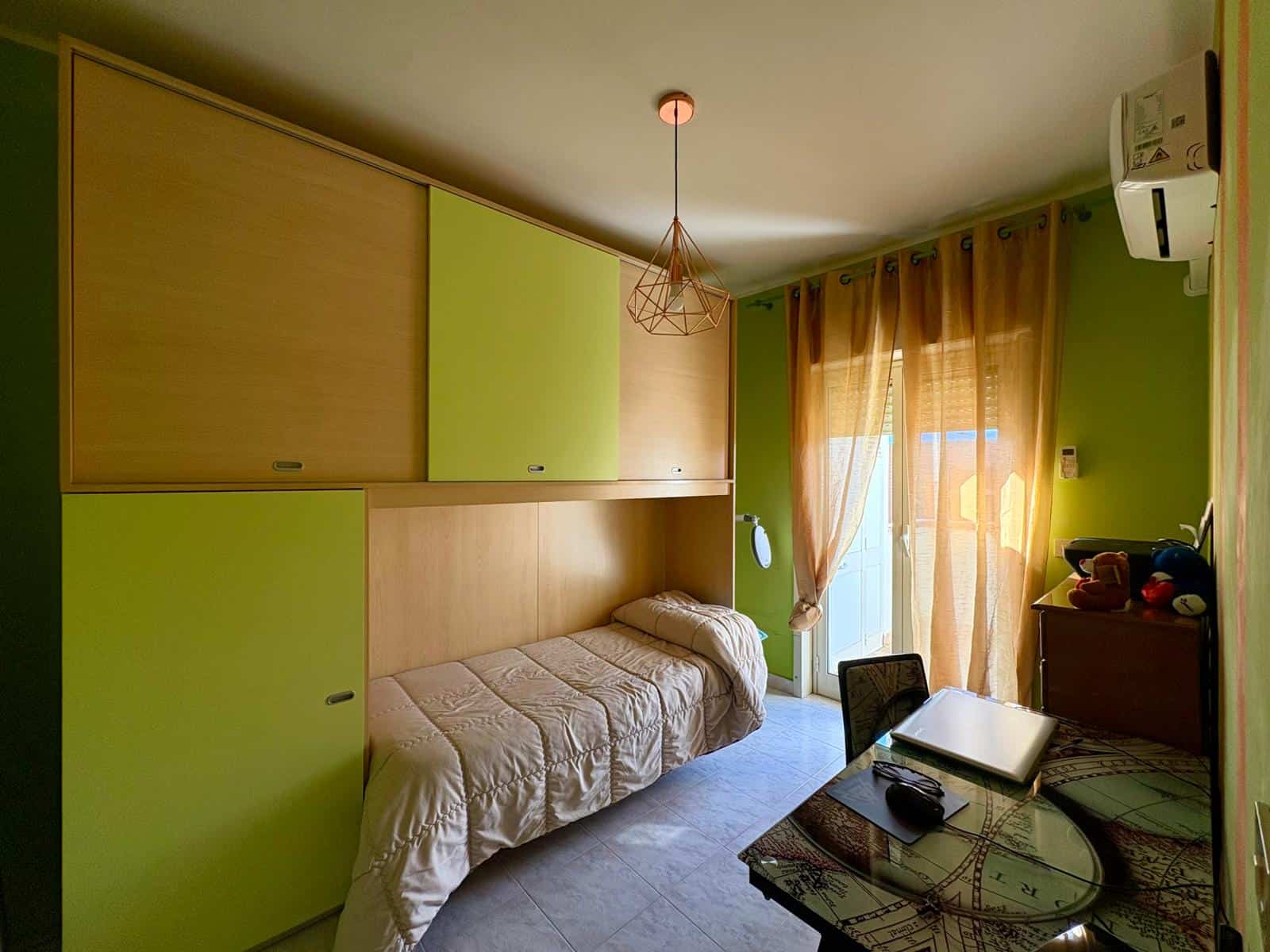 Letto singolo in camera con armadio a ponte in verde e legno, ambiente luminoso con tende beige e aria condizionata. Ideale per soluzioni di affitto breve o residenziali.