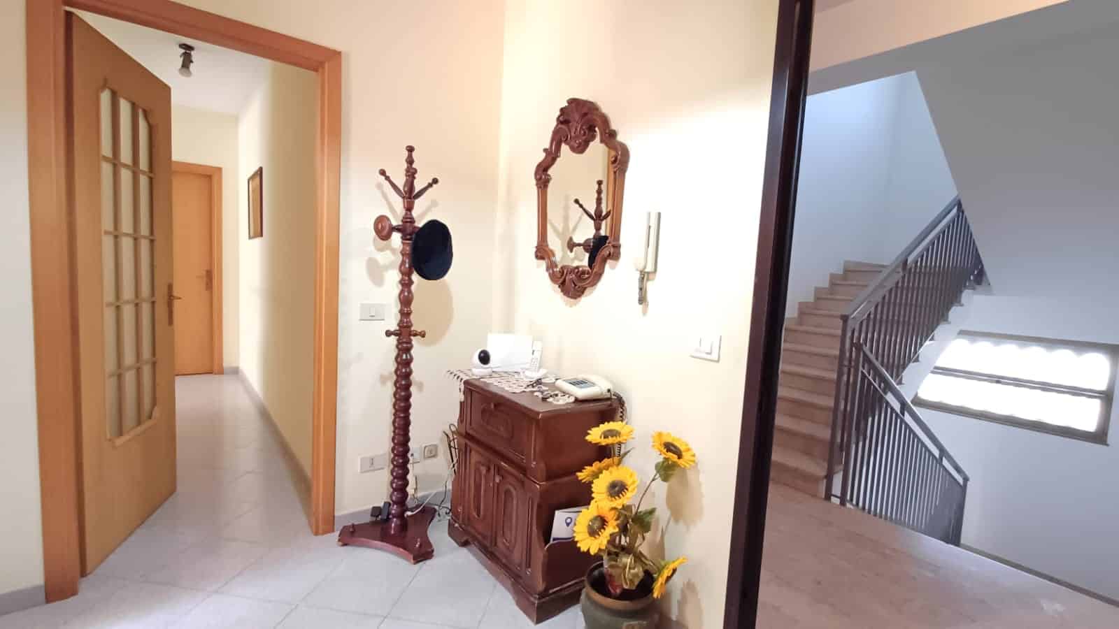 1. corridoio ingresso casa con mobili in stile, specchio, appendiabiti, vaso con girasoli, scale interne e lampada a soffitto.