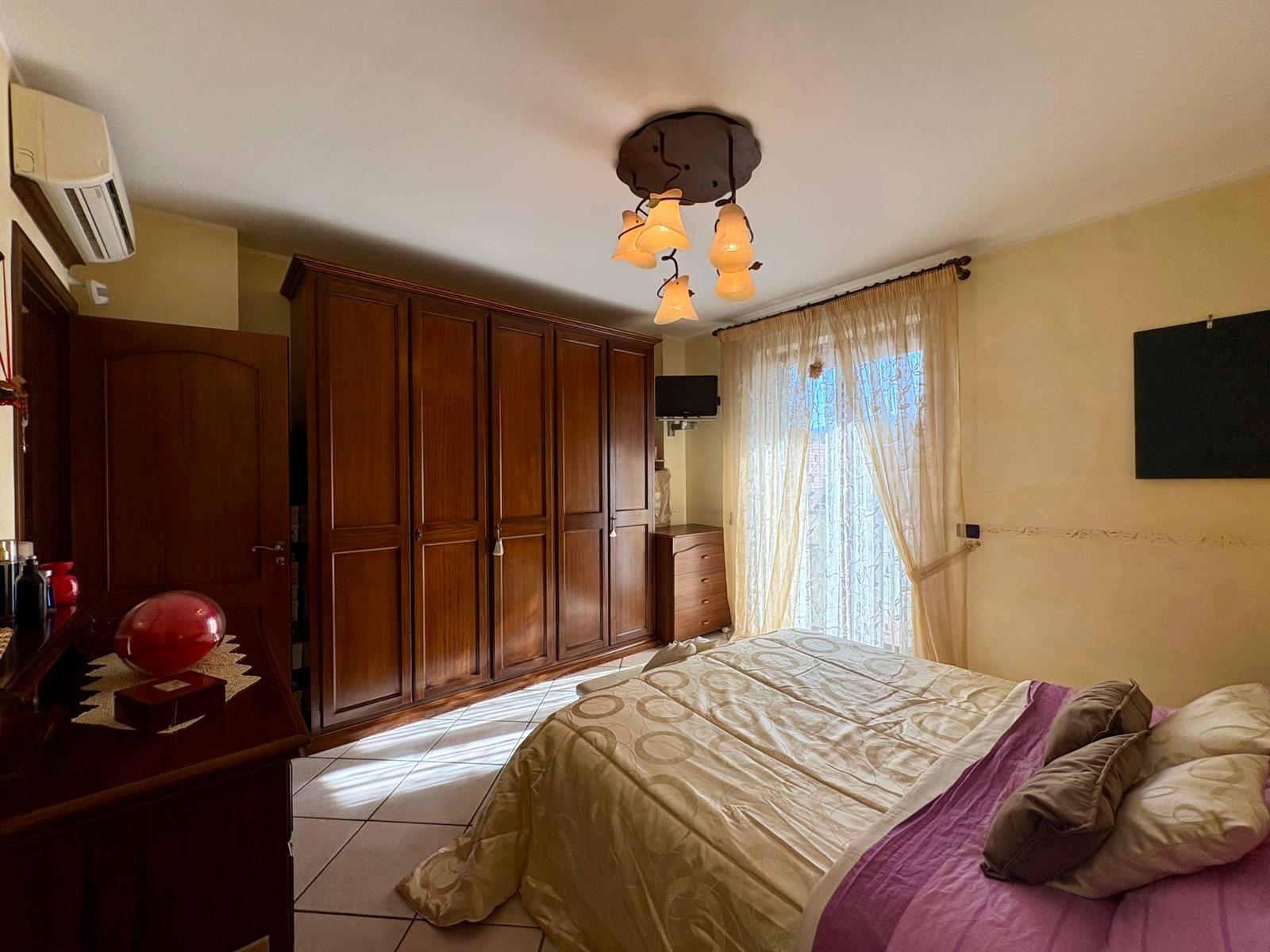 Elegante camera da letto con grande armadio in legno, letto matrimoniale con lenzuola beige e cuscini, finestra con tende leggere, aria condizionata e TV a schermo piatto.