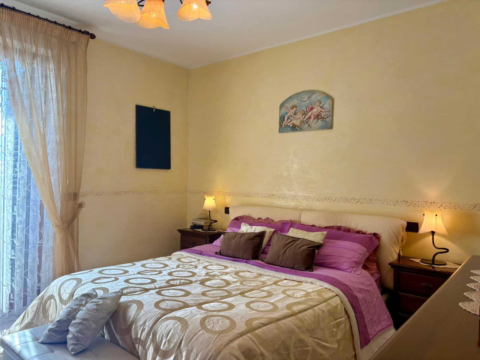 Ampia camera matrimoniale con letto king size, arredamento elegante, illuminazione calda, tende leggere, decorazioni artistiche e atmosfera accogliente per immobili in vendita o affitto.
