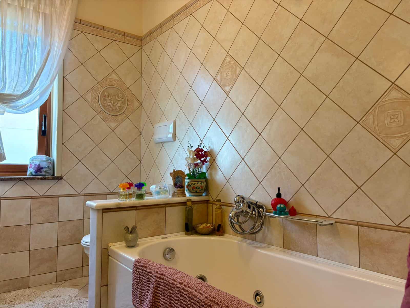 Bagno con vasca idromassaggio, arredato con dettagli decorativi e piante, finestra grande con tendina, ambiente accogliente e luminoso, ideale per relax e benessere domestico.