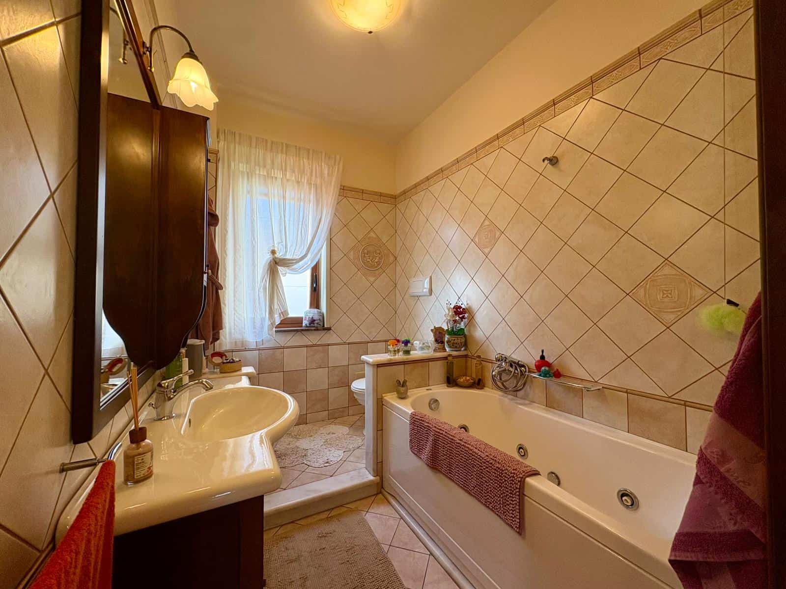Bagno spazioso con vasca idromassaggio, lavabo e specchio con illuminazione, finestra con tende leggere, decorazioni floreali e arredi in stile classico. Perfetto per relax e benessere domestico.